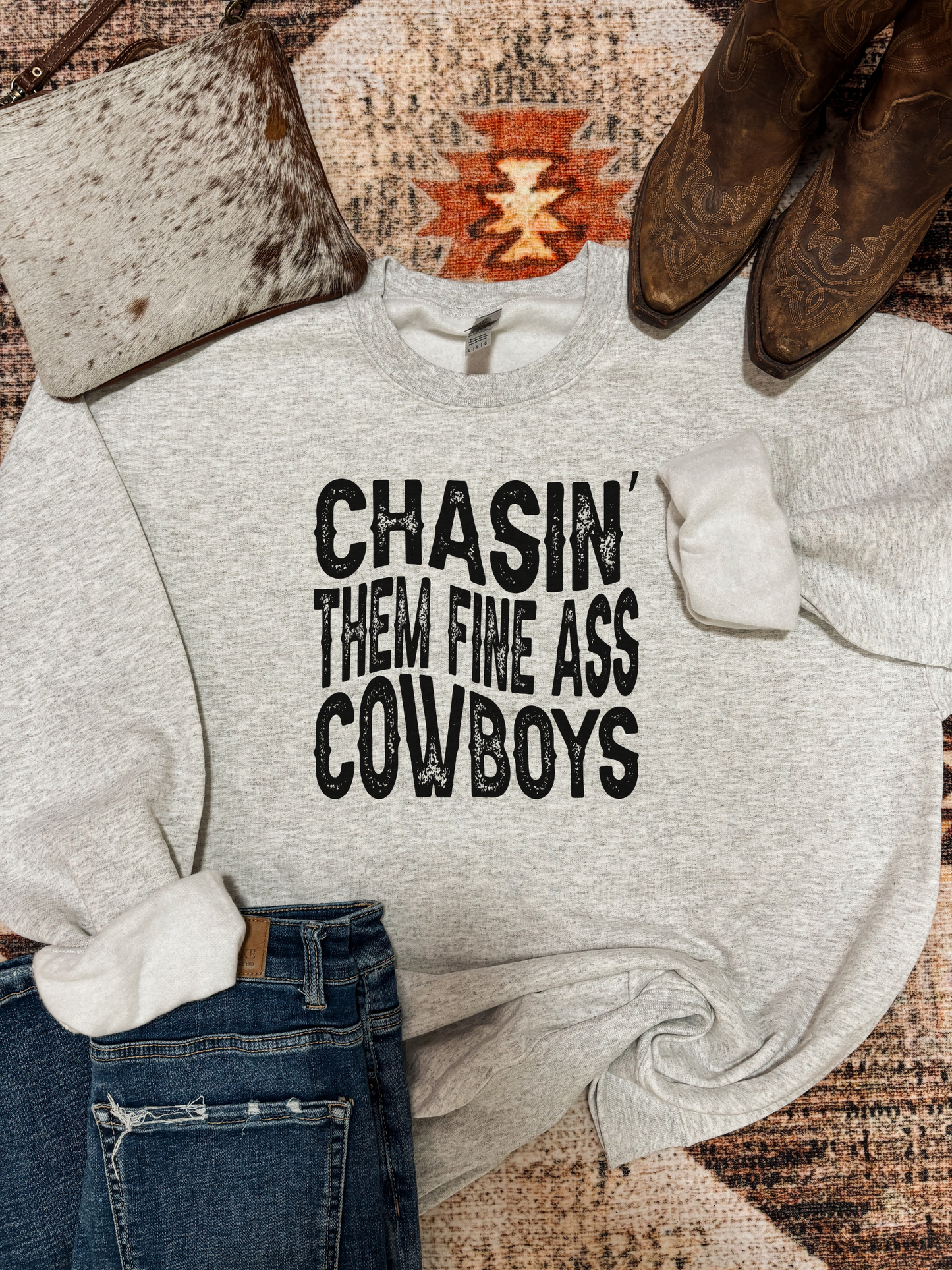 Heather grey crewneck with bold black western text that reads "Chasin' Them Fine Ass Cowboys," styled with jeans, cowboy boots, and a cowhide bag on a southwestern rug.