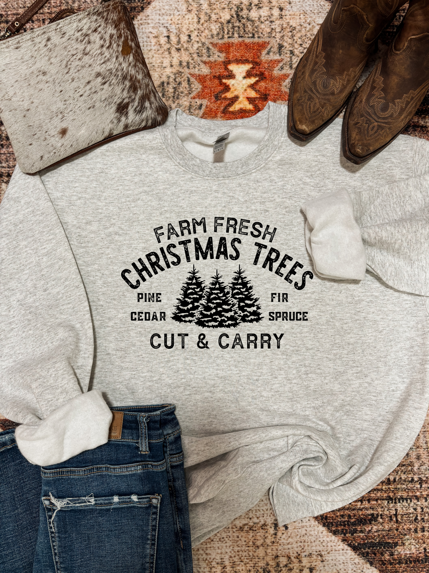Heather grey crewneck with a vintage-style black graphic that reads “Farm Fresh Christmas Trees – Pine, Fir, Cedar, Spruce – Cut & Carry,” styled with jeans, boots, and a cowhide bag on a rustic rug.