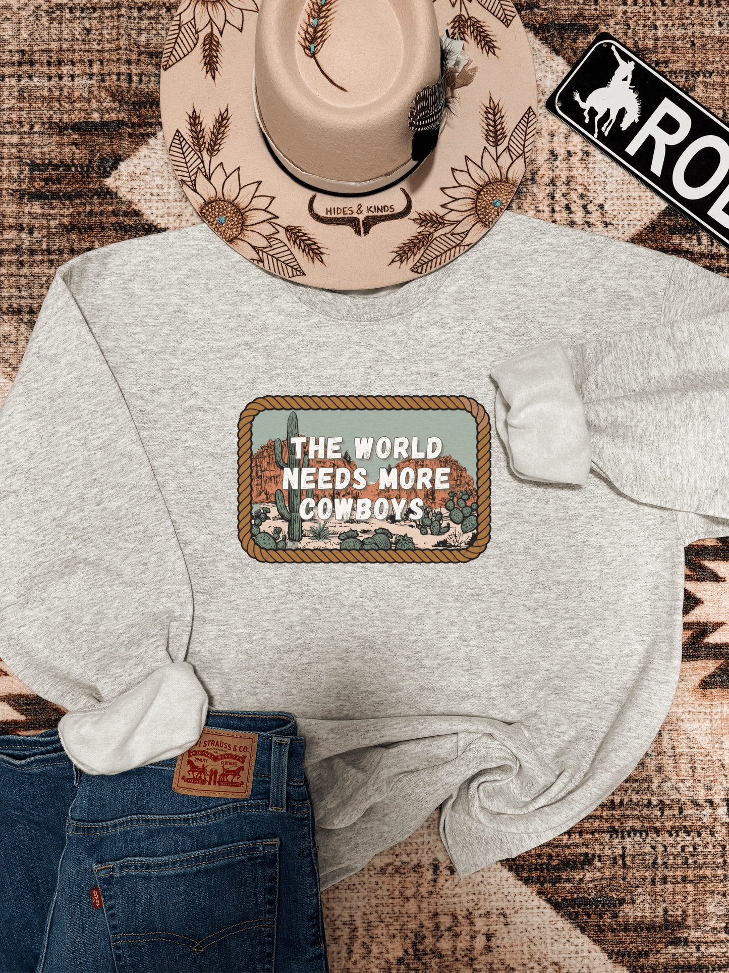 Heather grey crewneck laid flat with "The World Needs More Cowboys" graphic and desert cactus scene