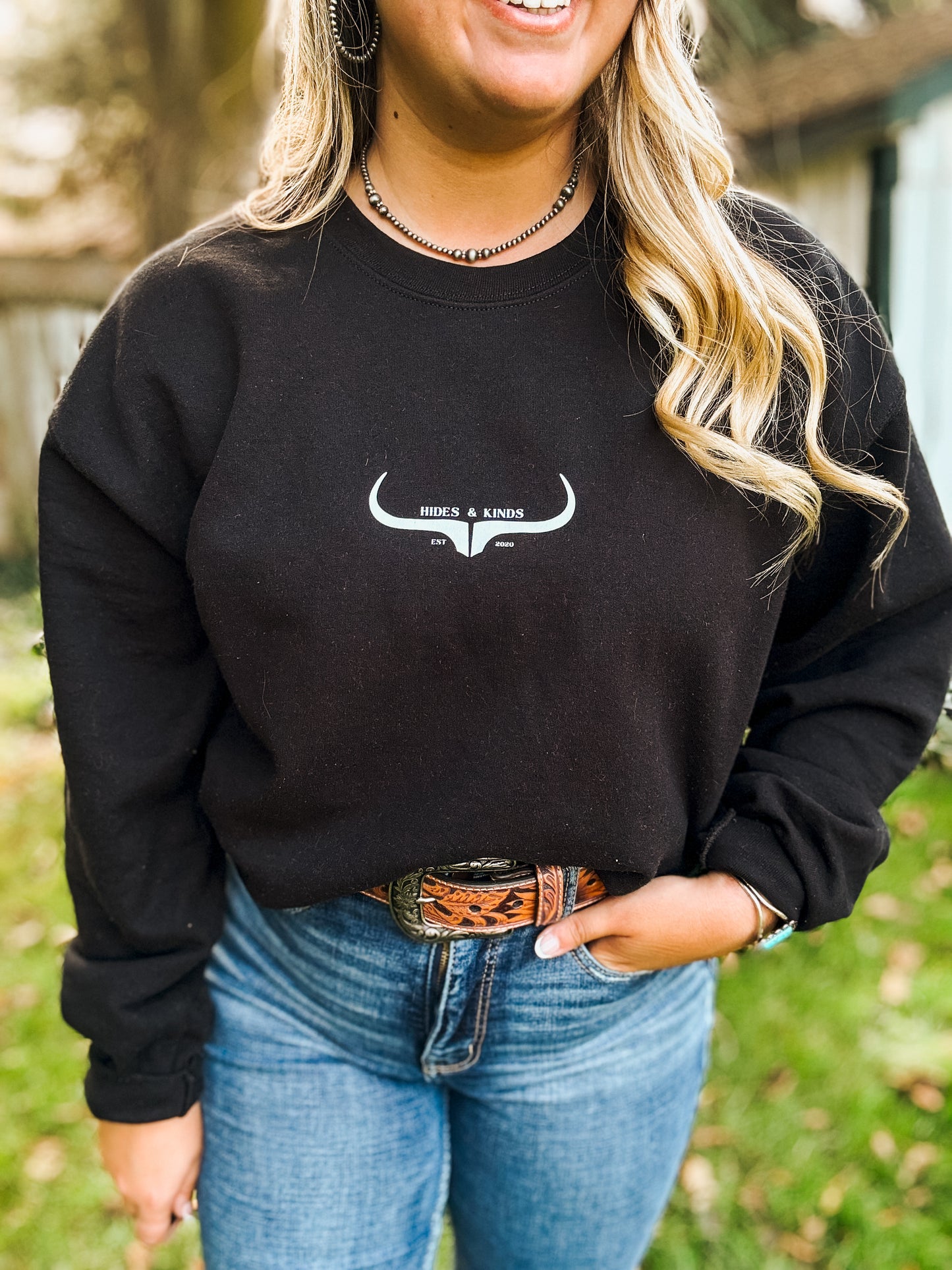 Woman wearing black Hides & Kinds crewneck with white longhorn logo, paired with a western tooled leather belt and jeans , casual outdoor lifestyle look.