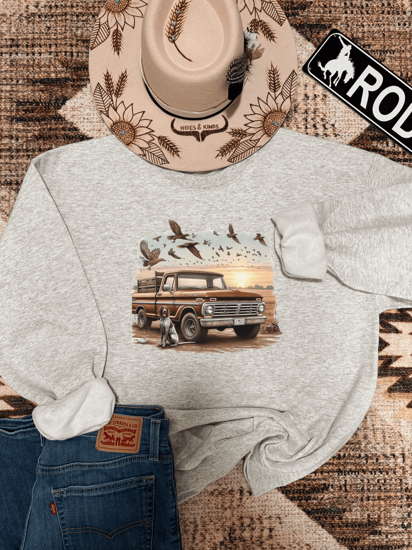 Grey crewneck with vintage truck and hunting dog graphic, styled with western hat, Levi’s jeans, and a rustic rodeo sign on a boho rug