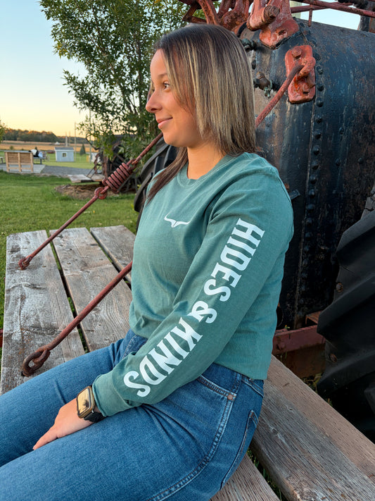 Model wearing the green Hides & Kinds Branded Longsleeve with white lettering down the sleeve and a white longhorn chest logo, seated outdoors beside vintage farm machinery
