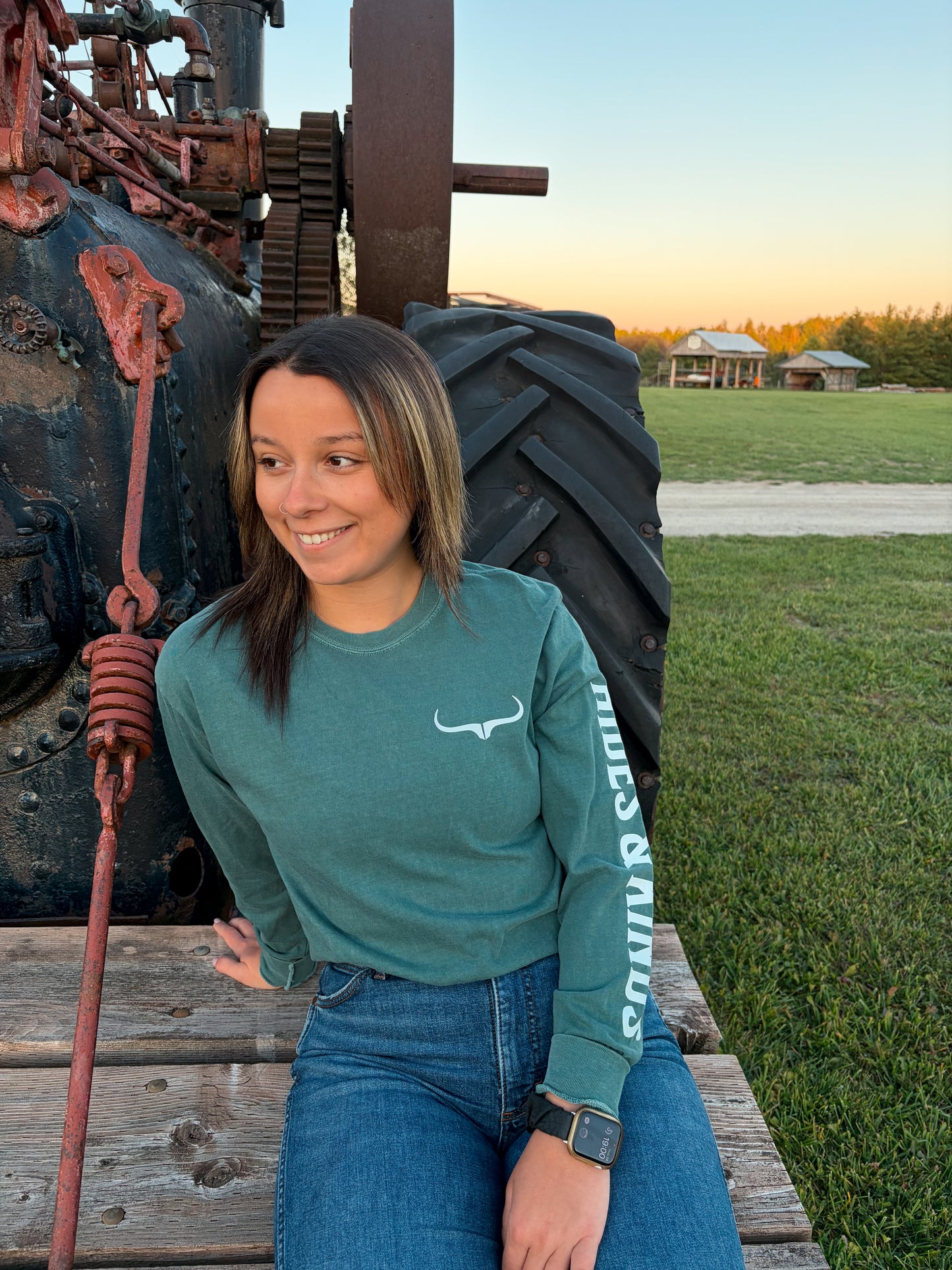 Front view of the green Branded Longsleeve by Hides & Kinds, showing the white longhorn chest logo and Hides & Kinds sleeve lettering, styled with blue jeans.