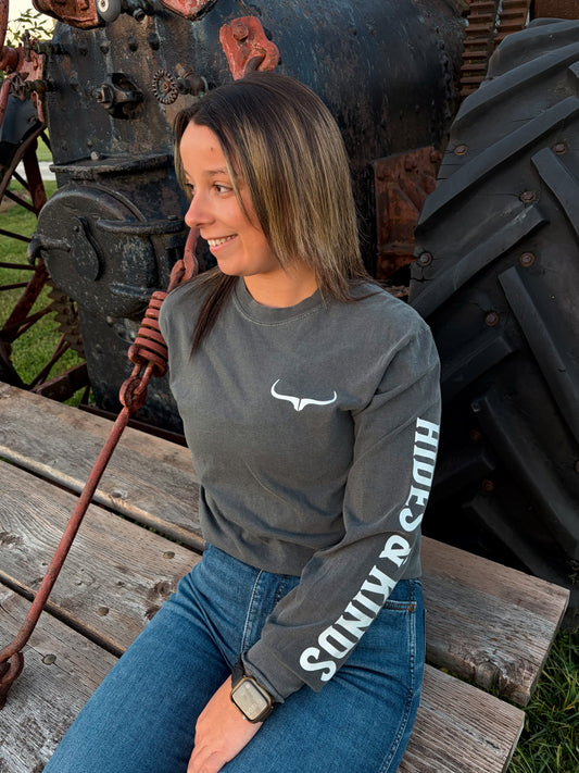 Front three-quarter view of the pepper grey Branded Longsleeve by Hides & Kinds, showing the white longhorn chest logo and bold Hides & Kinds sleeve lettering, styled with blue jeans.