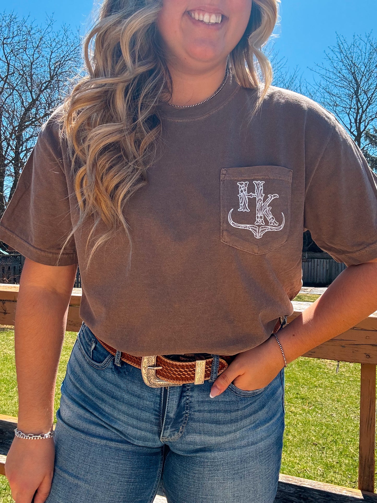 Detailed view of the espresso brown pocket tee with HK logo, styled casually with jeans and a western belt.