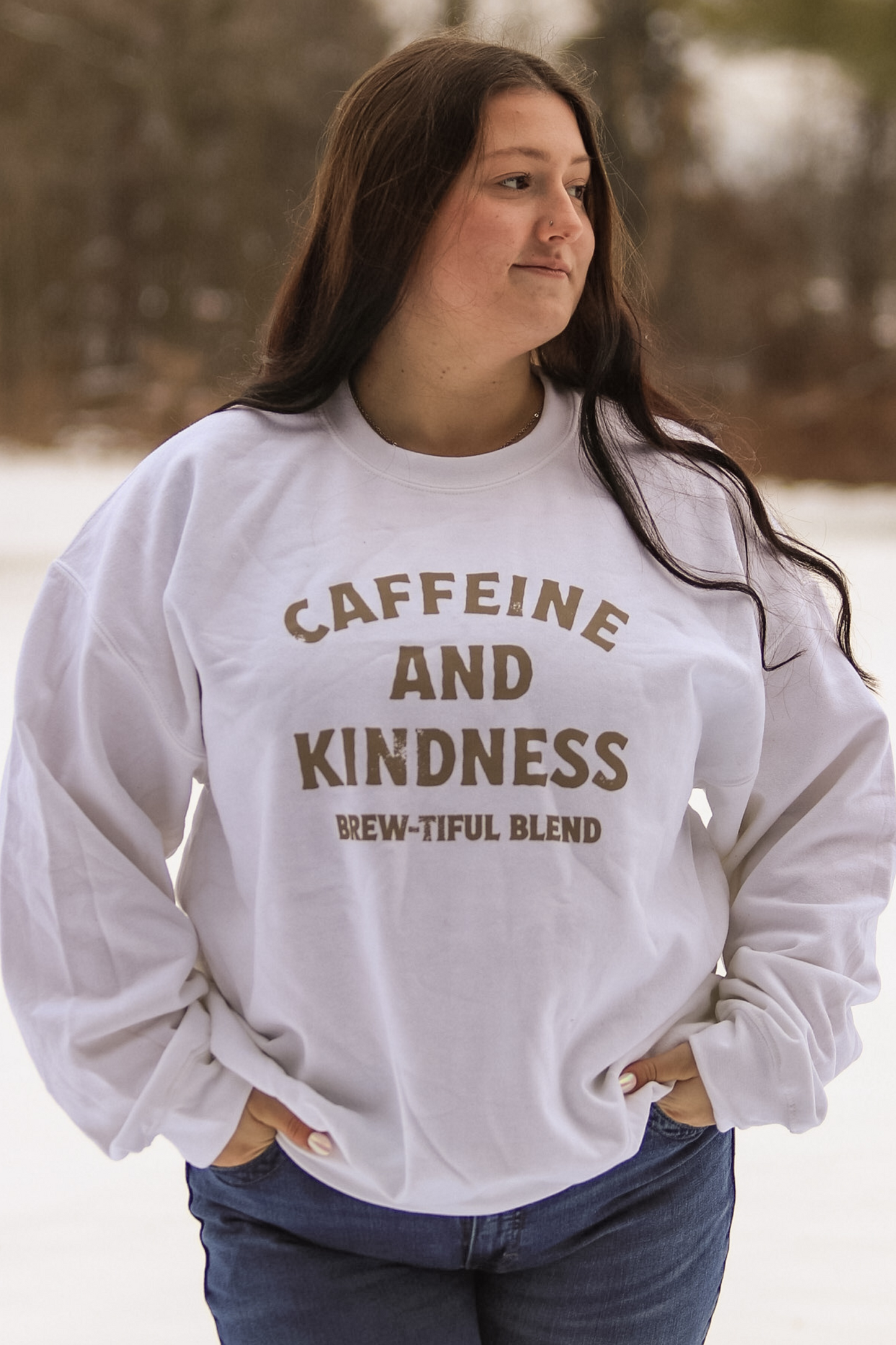 Close up of a white crewneck that says "Caffeine and Kindness Brew-tiful Blend" from Hides & Kinds, styled for winter.
