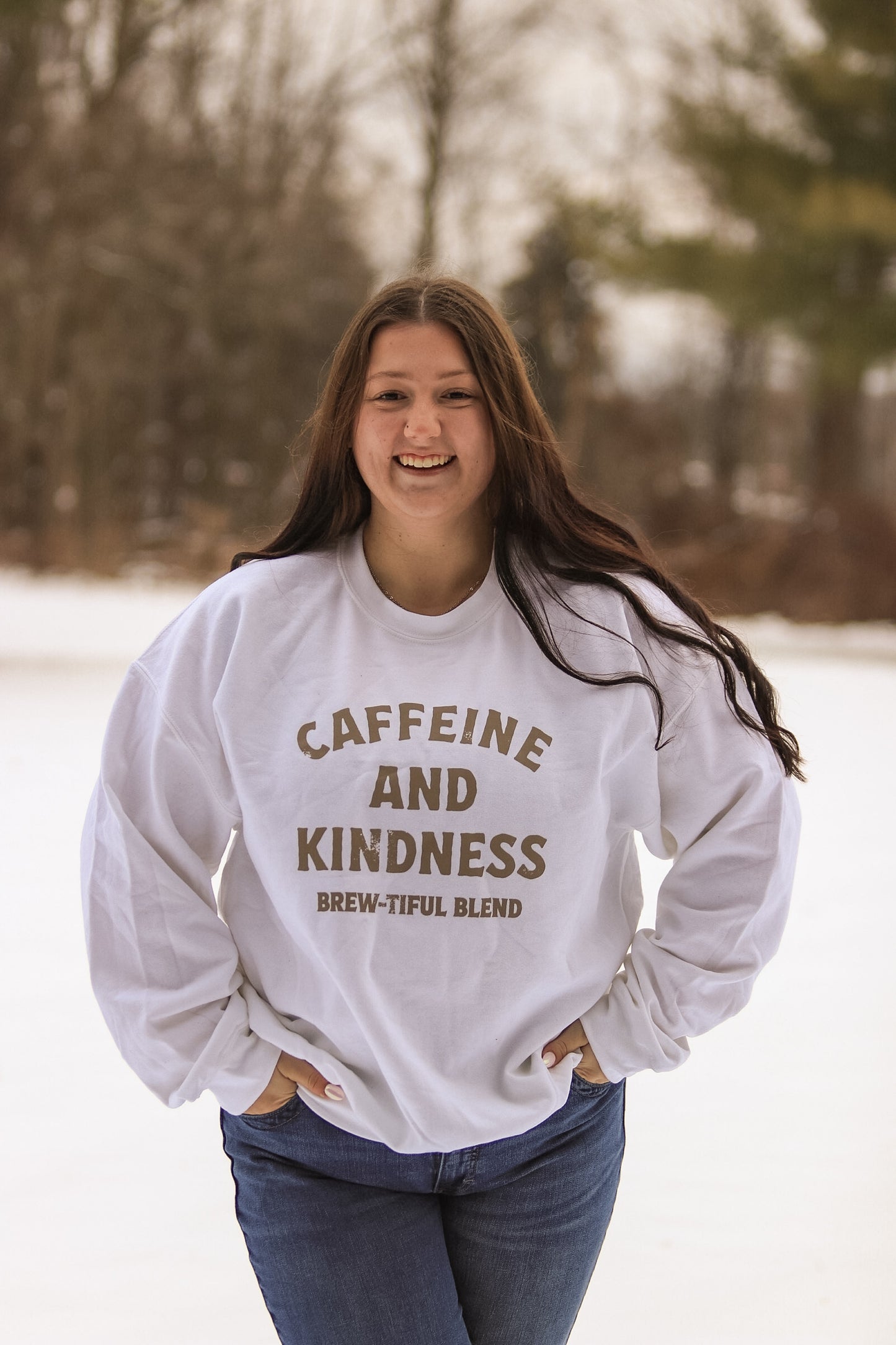 Woman smiling in the snow while wearing the Caffeine and Kindness crewneck sweatshirt by Hides & Kinds.