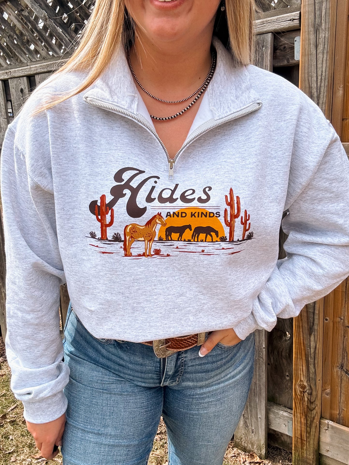 Full shot of model in Hides and Kinds Desert Horse Quarter Zip, paired with jeans and a western belt, standing by a rustic fence.