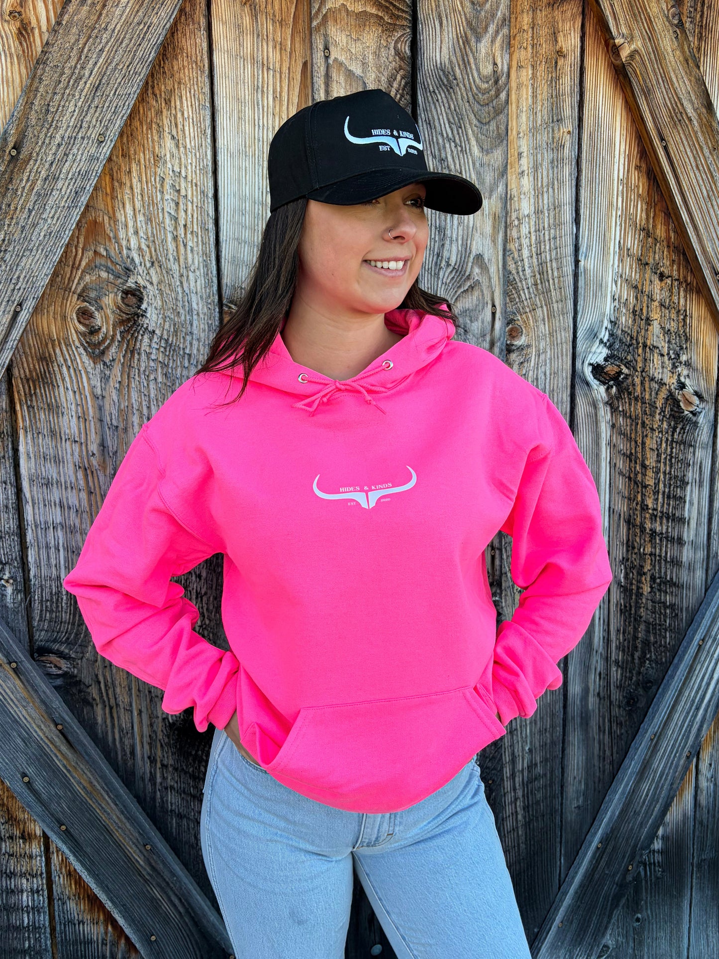 Neon pink Hides and Kinds hoodie with longhorn logo styled with black trucker hat and denim jeans