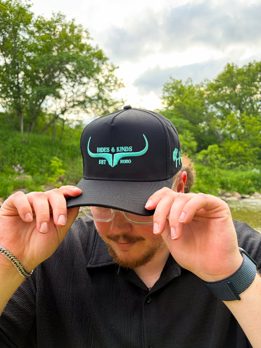 Man wearing the Hides & Kinds Original Logo Hat in black with teal longhorn embroidery