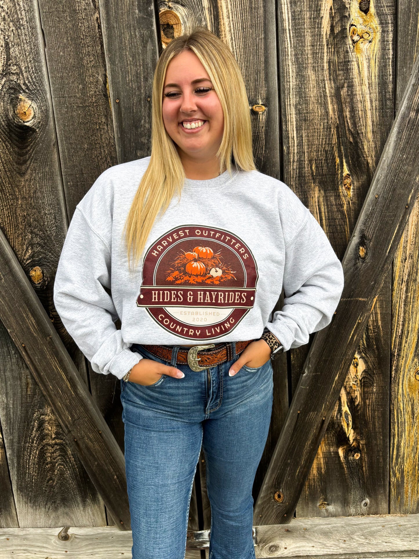 Smiling blonde woman wearing fall-themed Hides & Hayrides crewneck in grey with denim jeans and western belt