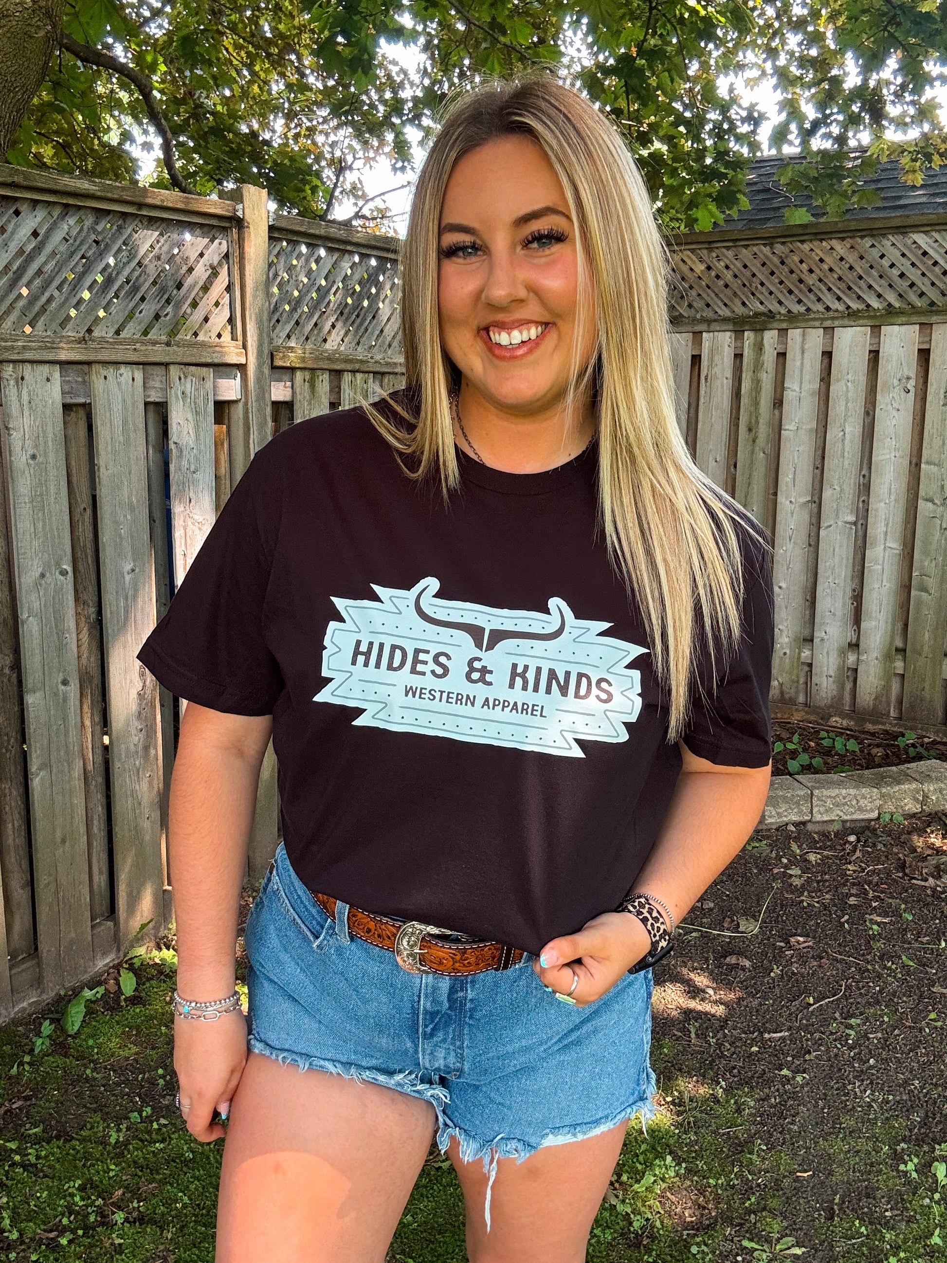 Woman wearing black Aztec Badge t-shirt from Hides and Kinds with turquoise logo, styled with denim shorts and a tooled leather belt.