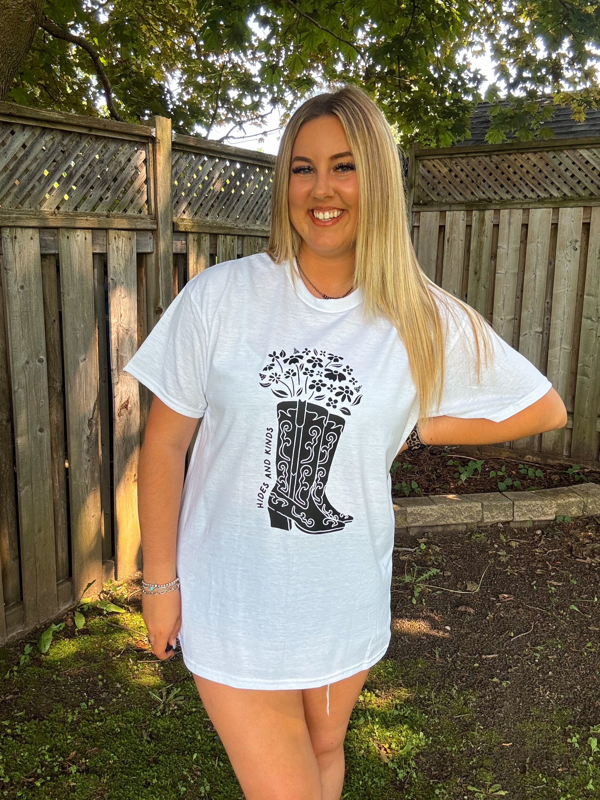 Smiling woman wearing Hides and Kinds floral cowboy boots t-shirt with black graphic design, outdoors.