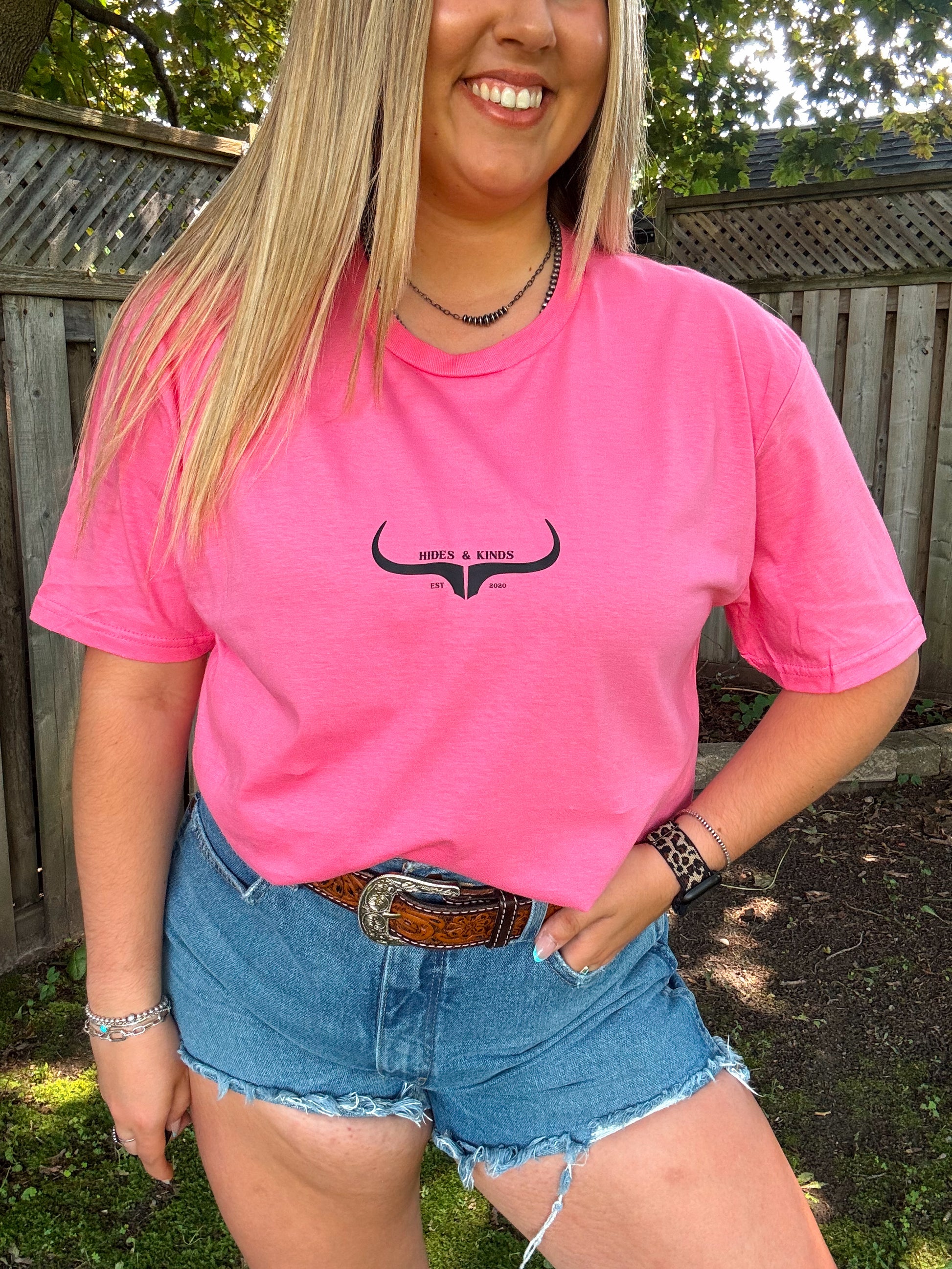 Close up of woman wearing pink Hides and Kinds logo tee styled with western belt and denim shorts.