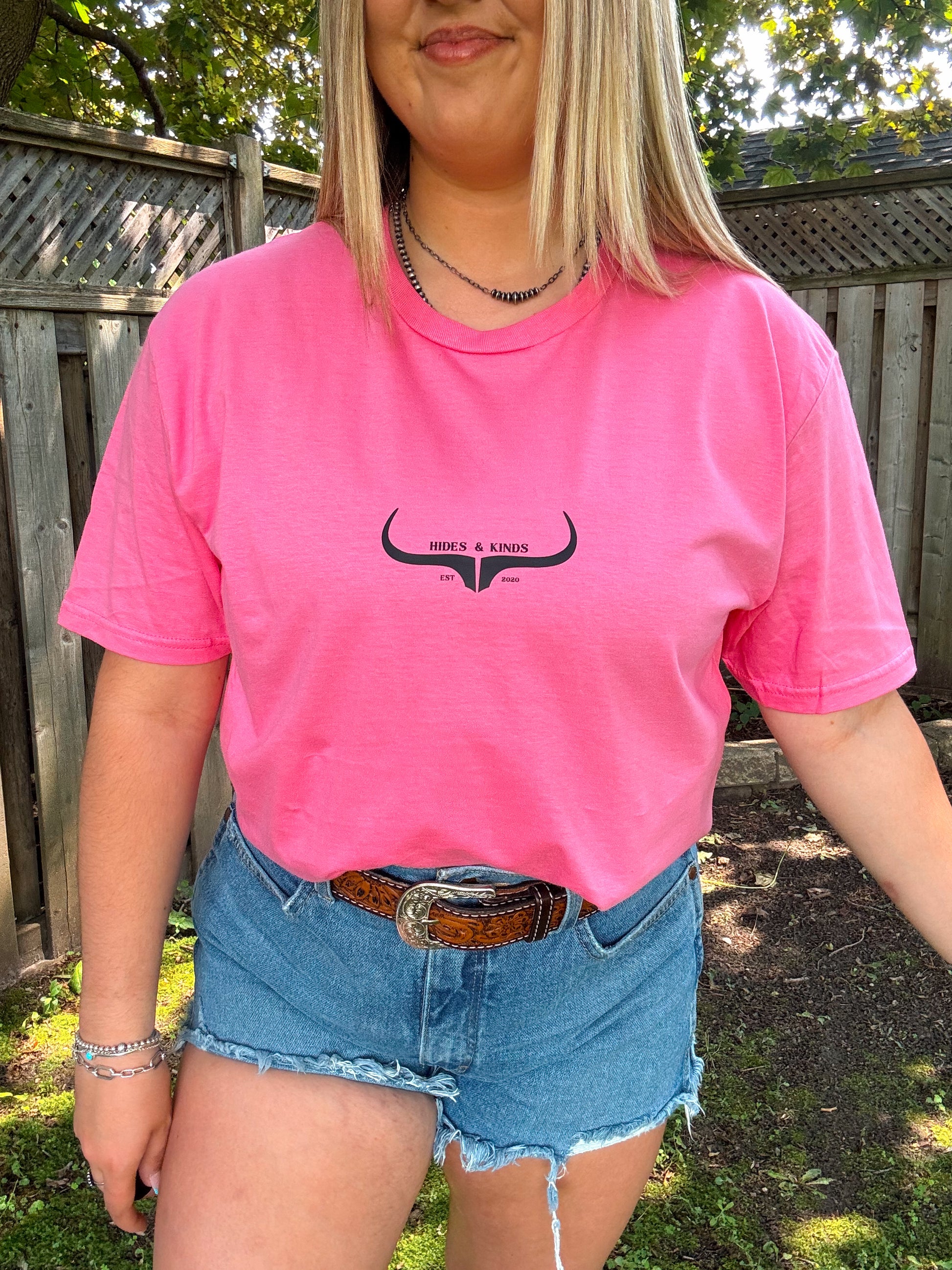 Front view of pink Hides and Kinds graphic tee styled casually with summer western accessories.