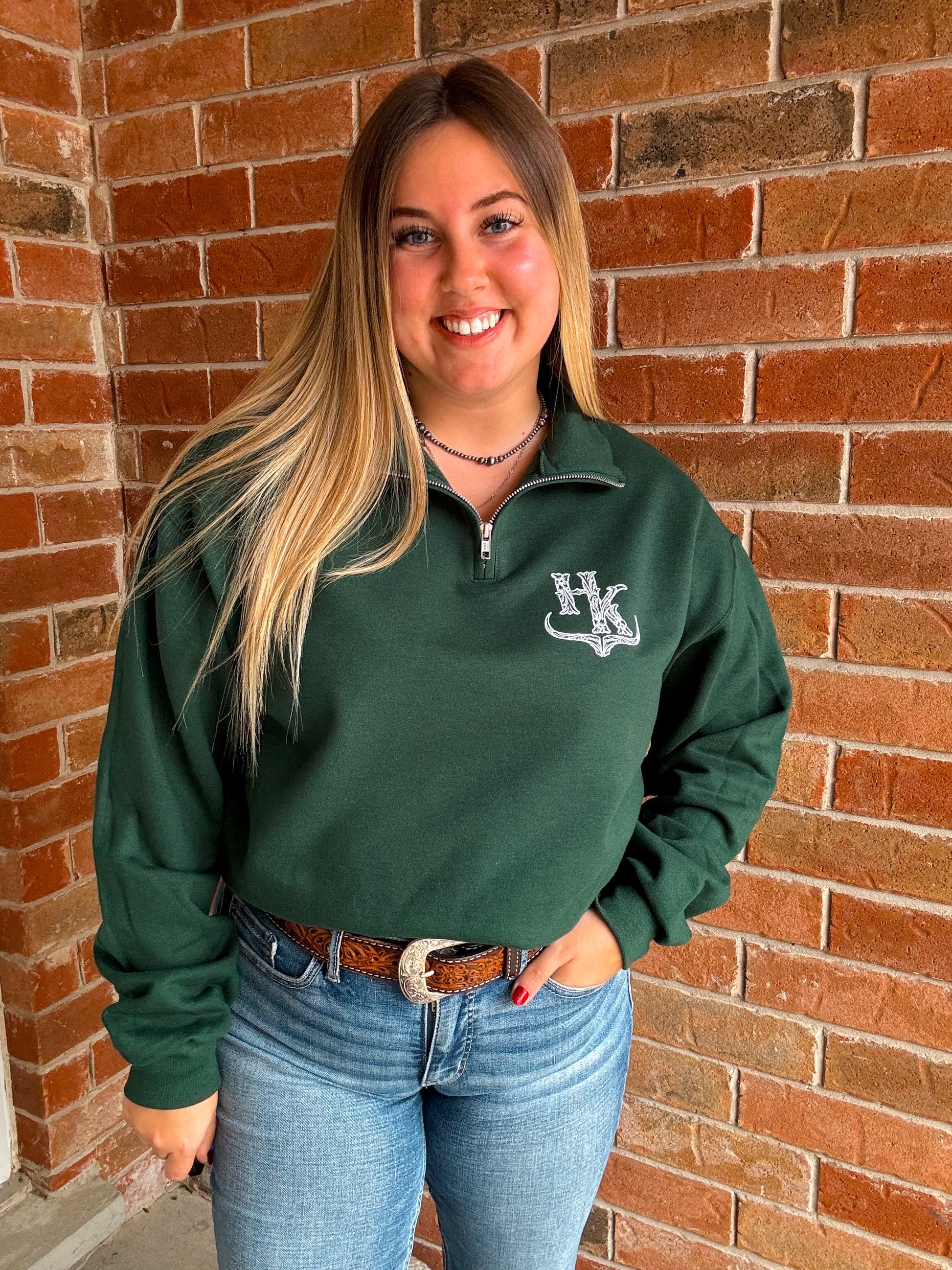 Full body shot of model in forest green Hides and Kinds quarter zip, styled western with a tooled belt and light wash jeans.