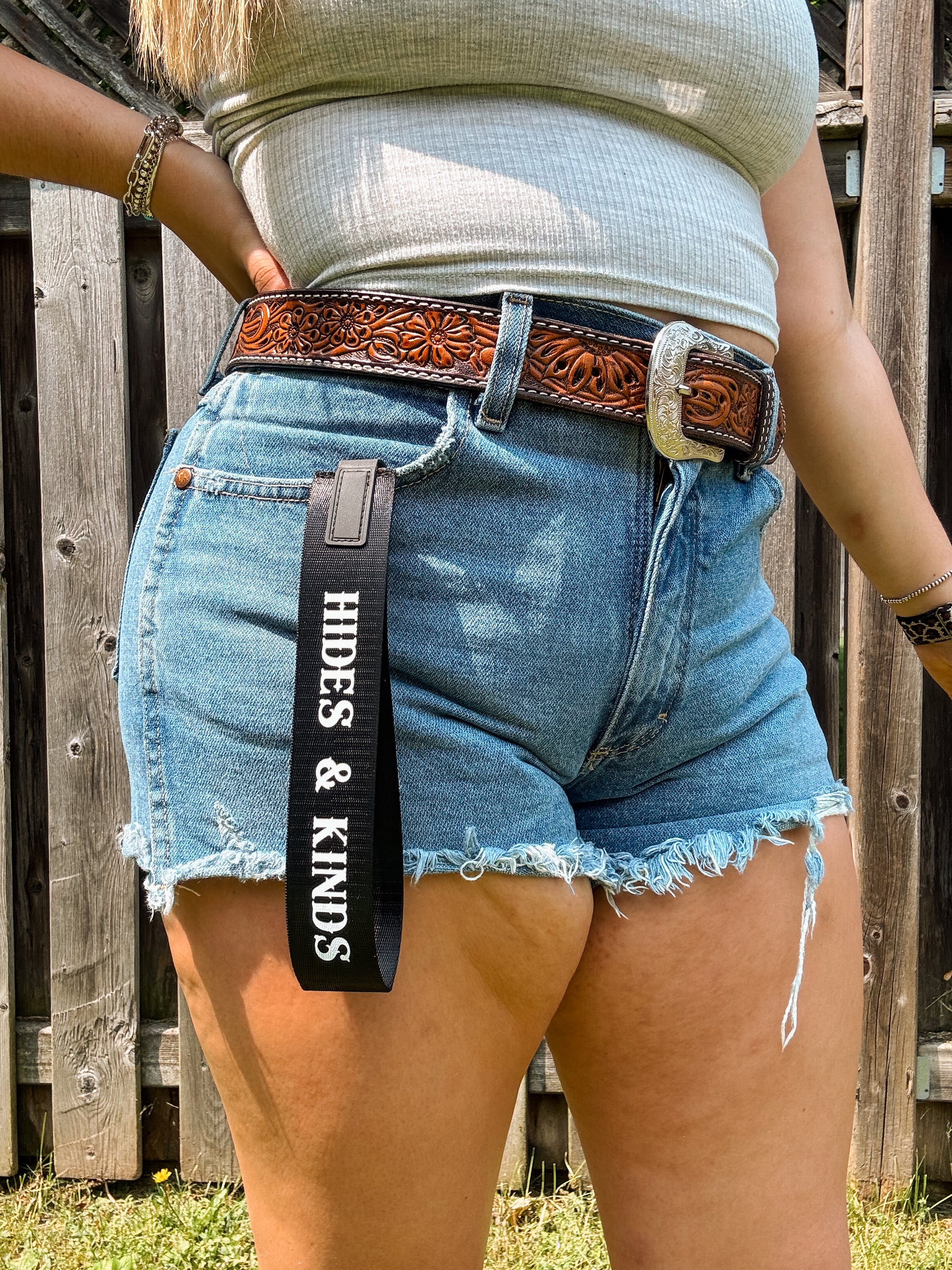 Black branded wristlet with white “Hides & Kinds” text, worn clipped to belt loop with western shorts and belt.
