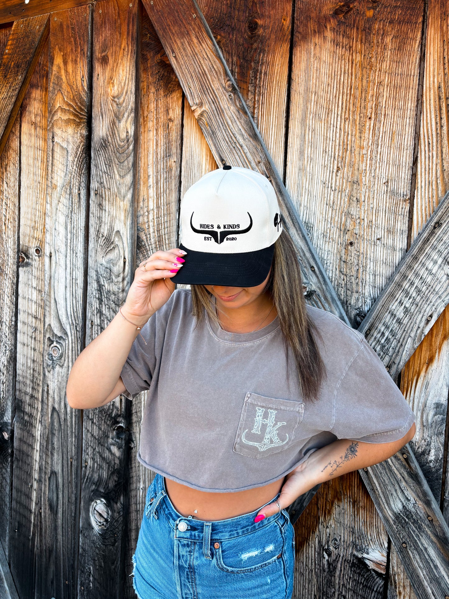 Model wearing the original Hides & Kinds logo hat paired with a cropped pocket tee and denim shorts in a rustic western setting.