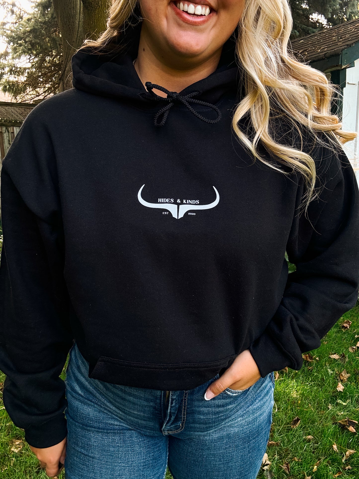 Woman wearing the Hides and Kinds Original Logo Hoodie in black with hands in pockets, showcasing western streetwear style outdoors