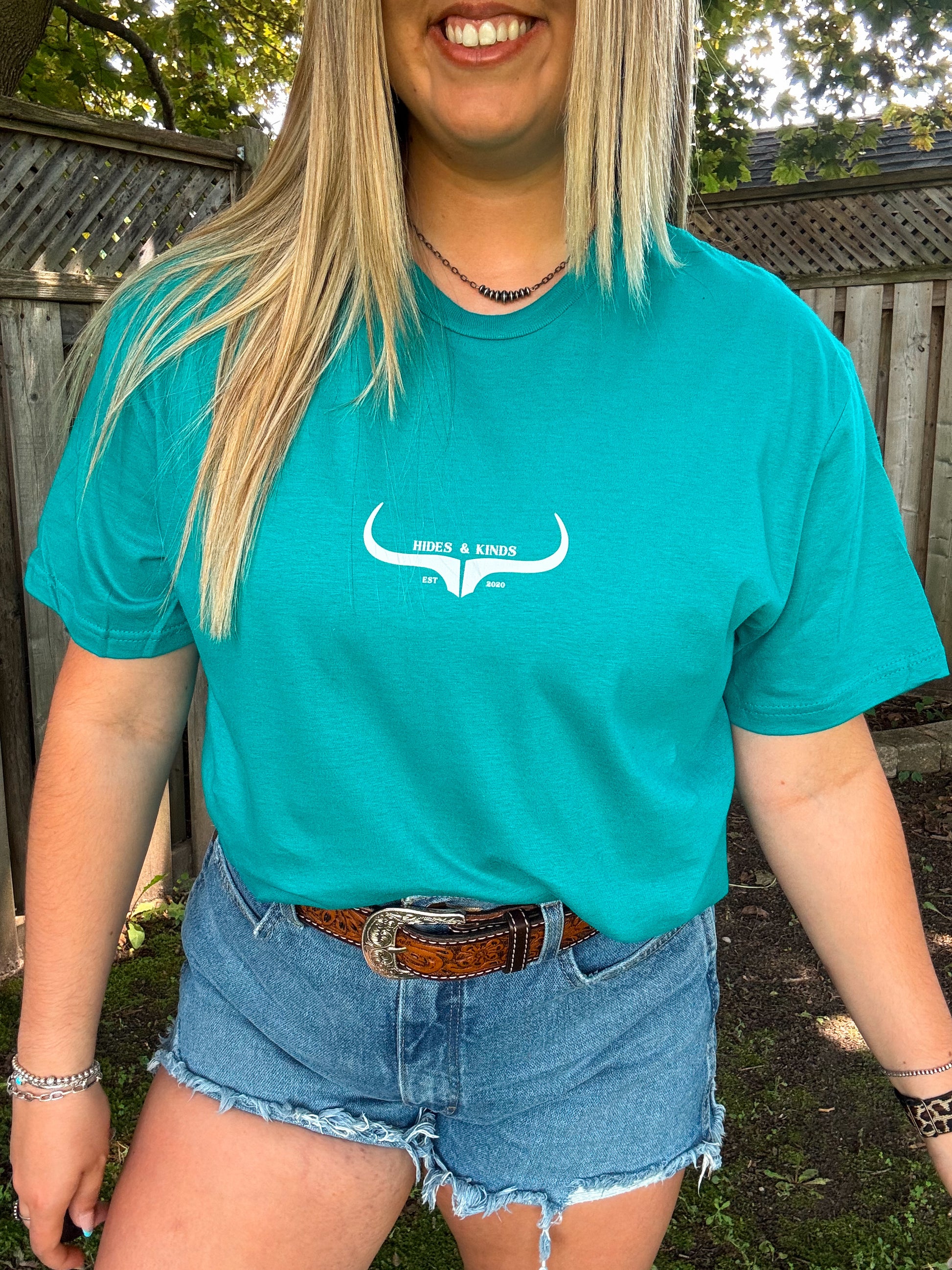 Close up of teal Hides and Kinds original logo t-shirt paired with denim shorts and silver accessories.