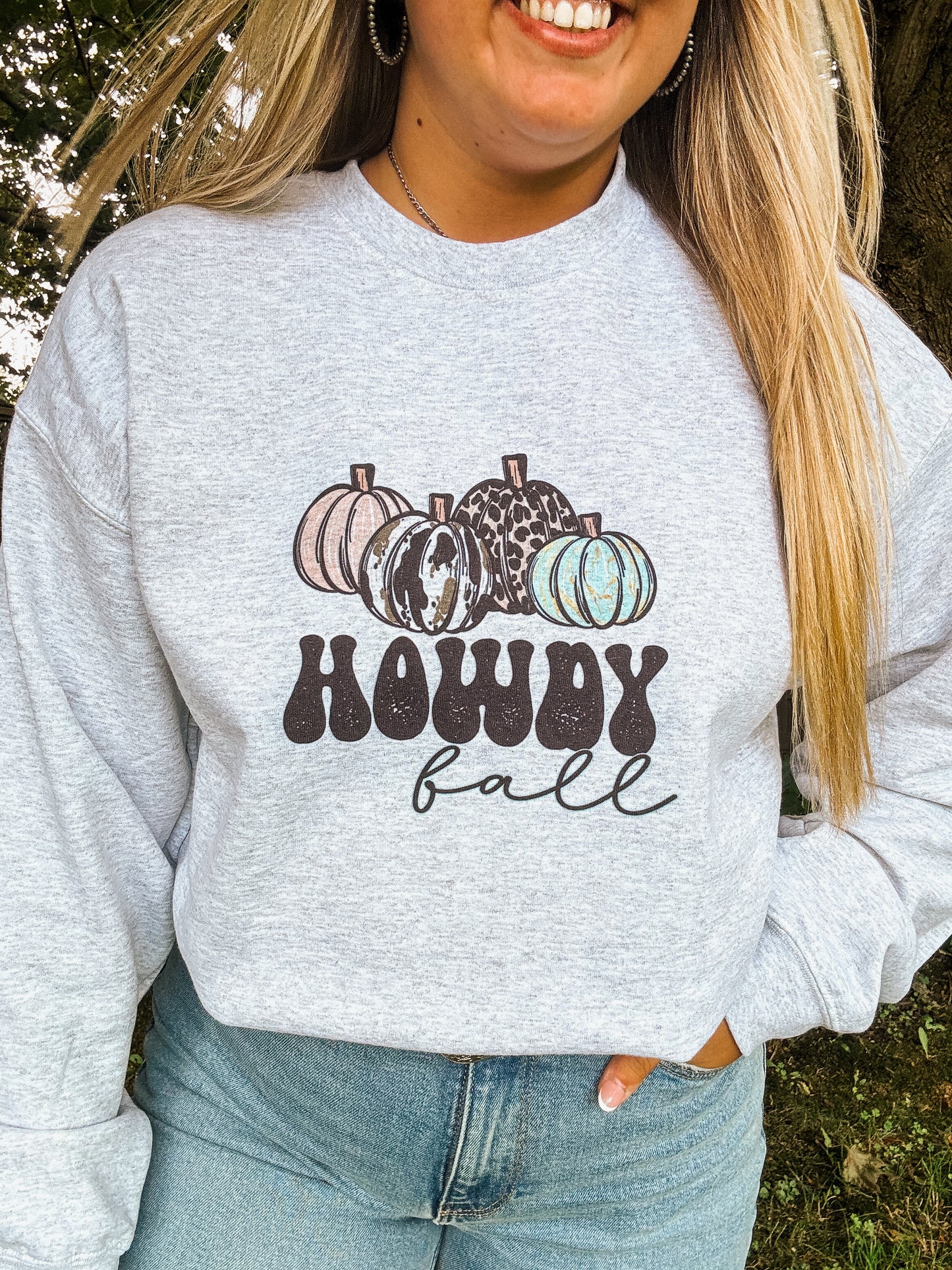 Woman wearing light grey crewneck with cow print and turquoise pumpkins and the text “Howdy Fall,” styled with denim jeans in outdoor setting
