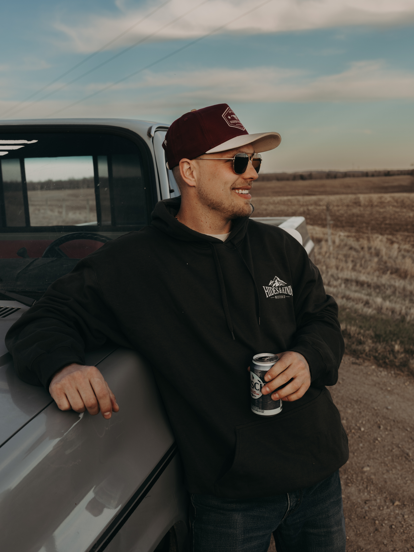 Man wearing the black Hides & Kinds Western Co hoodie with small chest logo, leaning against a vintage truck in a rural setting.
