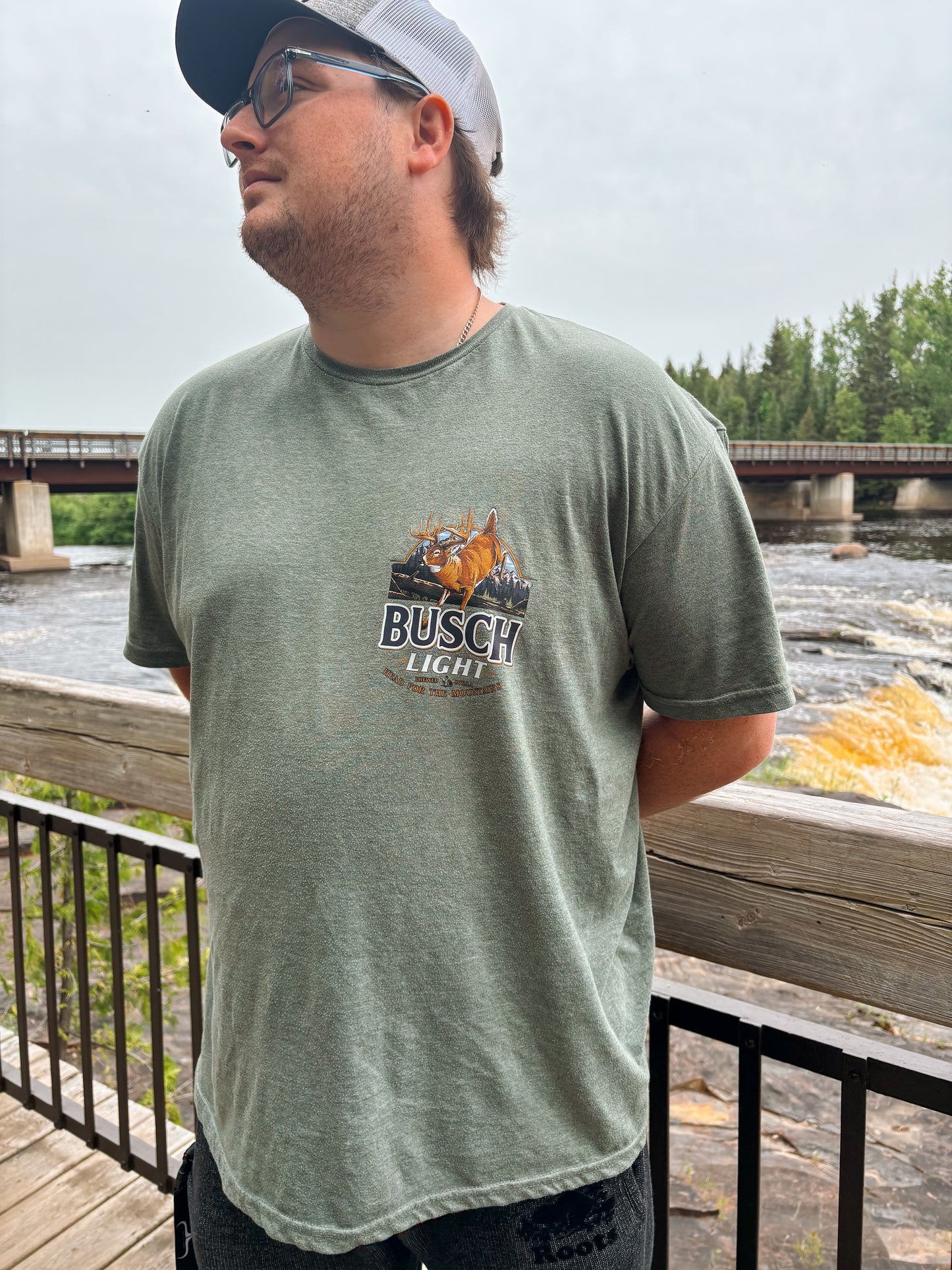 Man wearing the Mountain Buck Busch Tee with deer and mountain design on a sage green t-shirt near a river