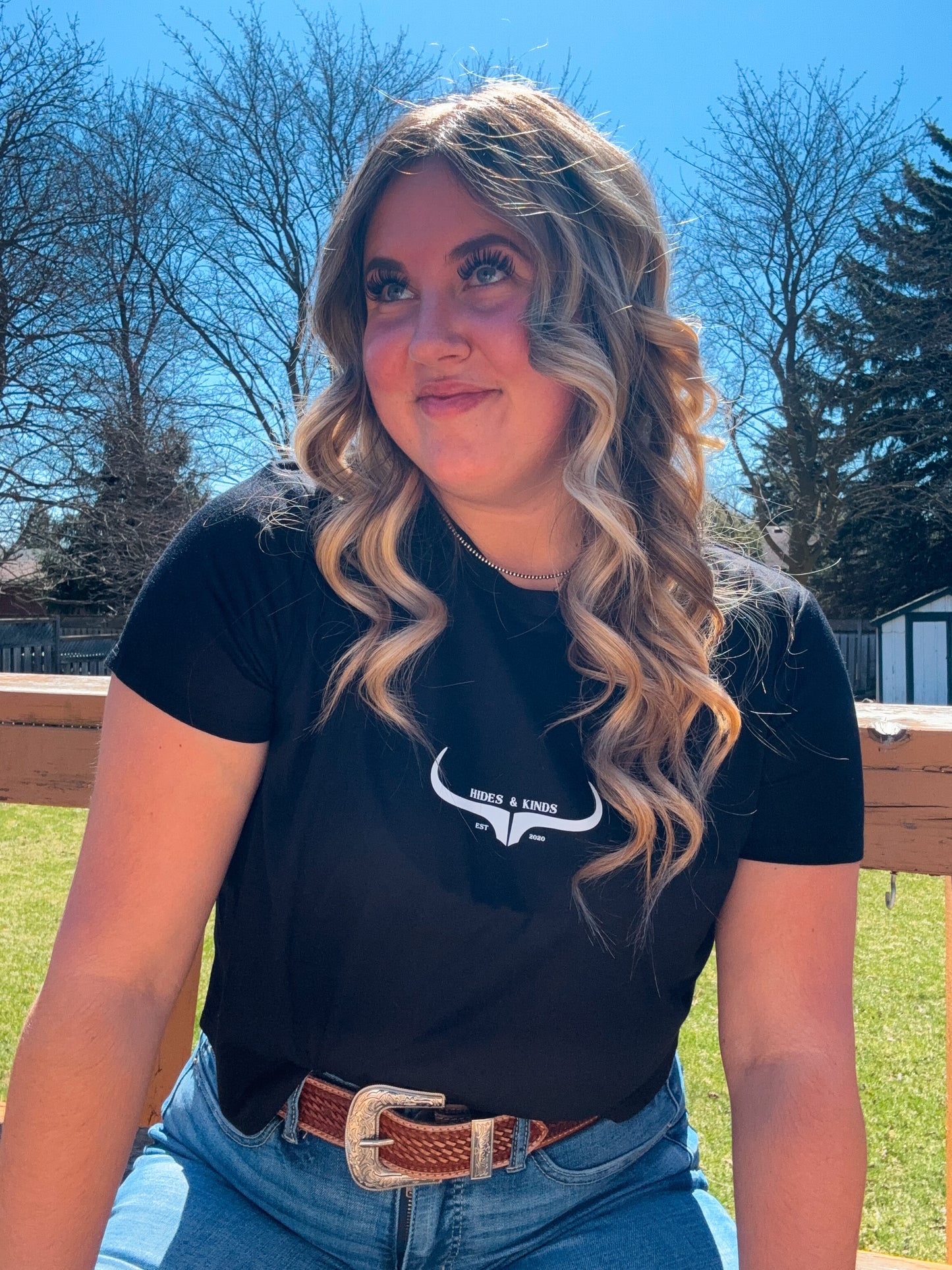 Model outdoors wearing the Hides and Kinds black cropped tee with curled hair and western belt, showing the tee in a natural setting.