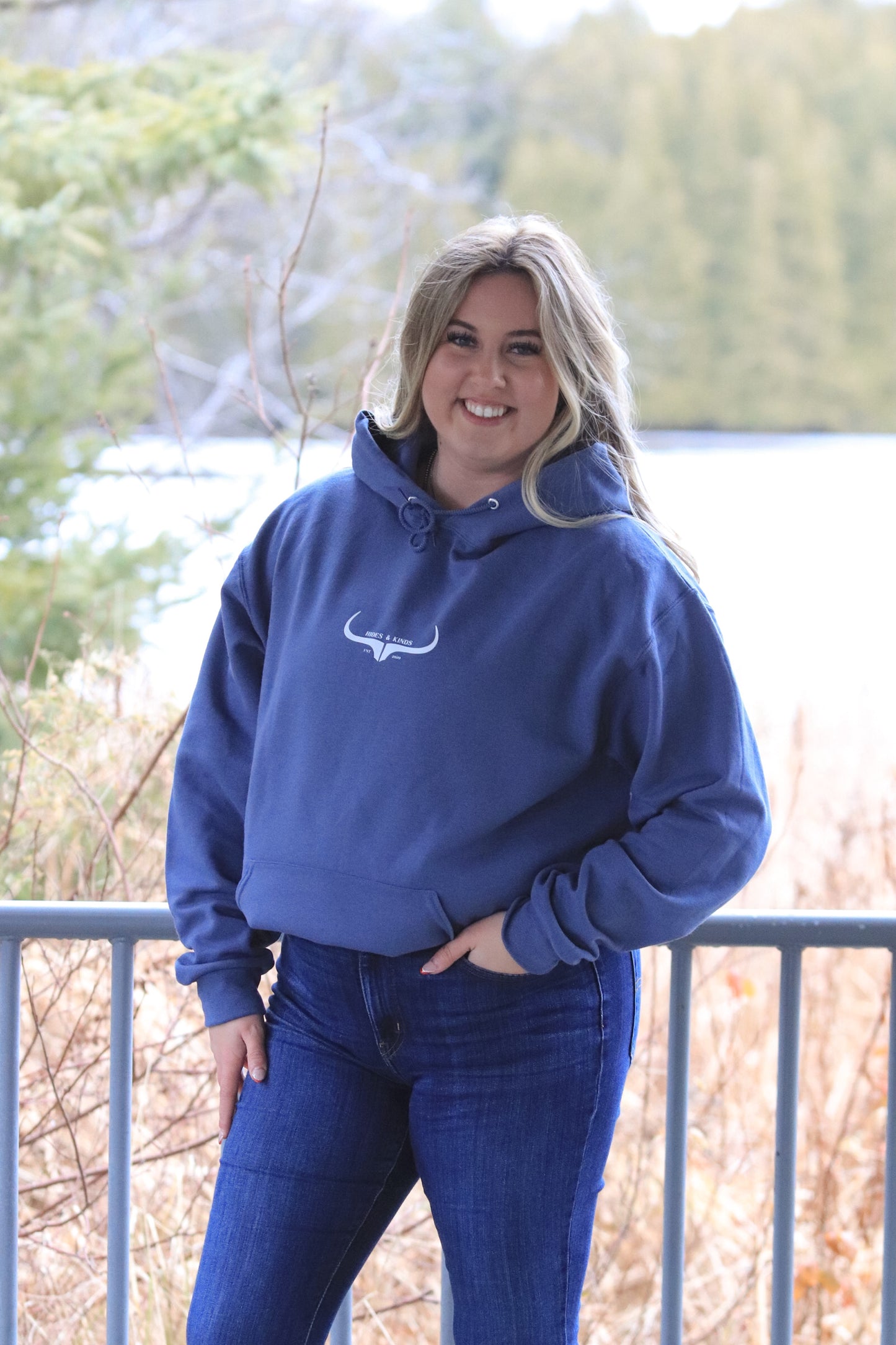 Full front facing view of a woman wearing the H&K Original Logo Hoodie in denim blue with natural forest scenery in the background.