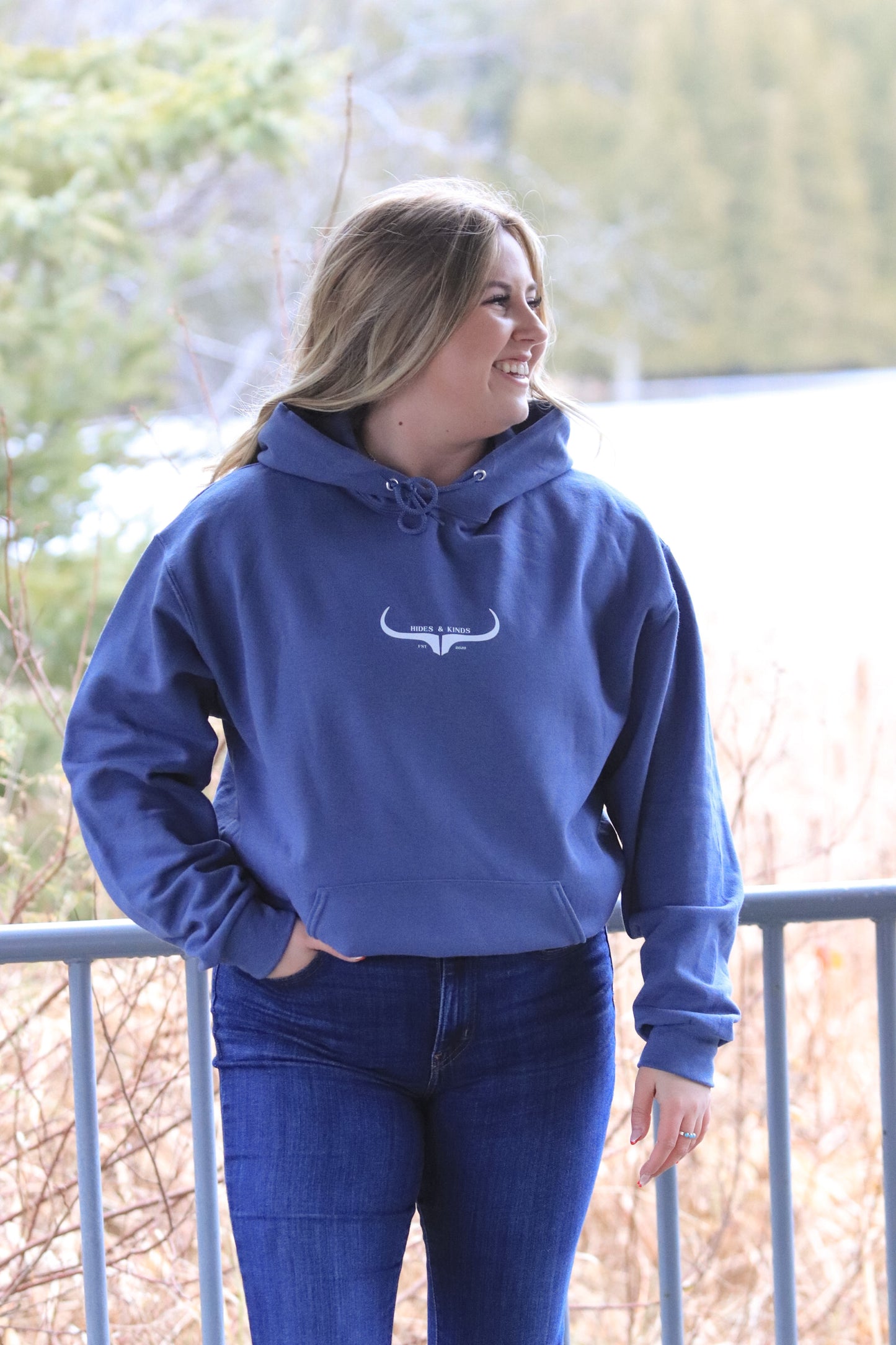 Woman smiling while wearing the Original Logo Hoodie in denim blue, styled casually outdoors with one hand in her pocket.