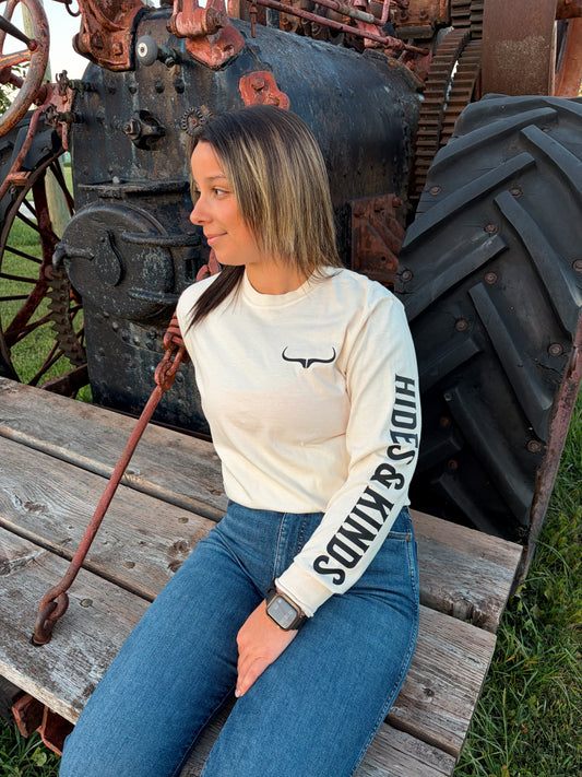 Front view of the cream Original Logo Long Sleeve by Hides & Kinds, showing the black longhorn chest logo and bold Hides & Kinds sleeve print, styled with blue jeans.