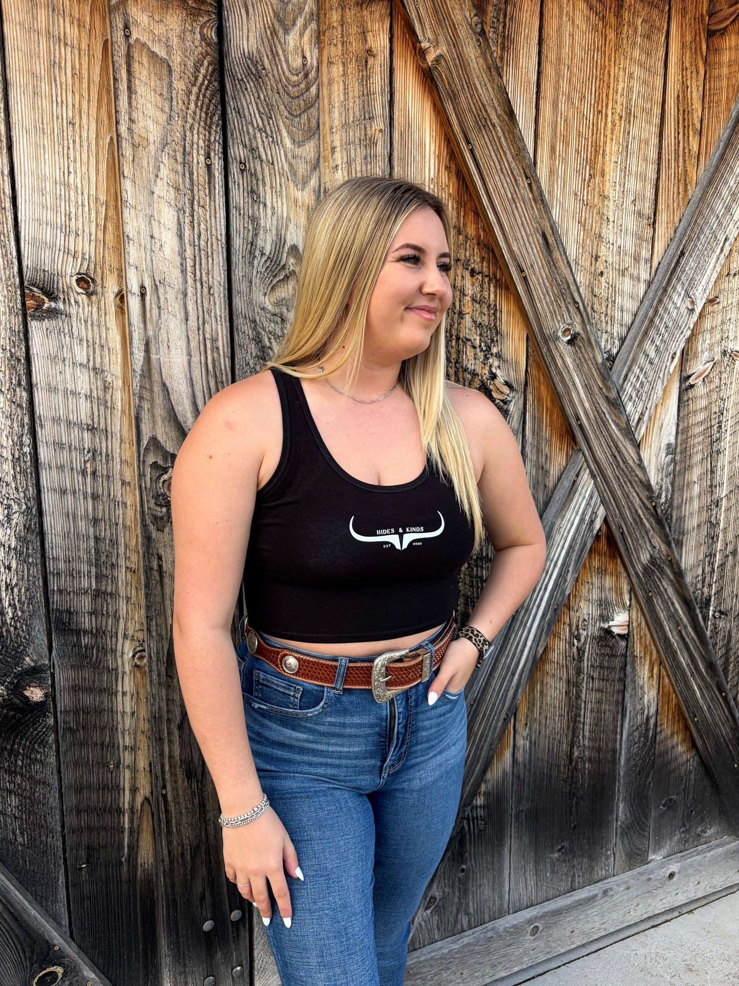 Black Hides & Kinds western cropped tank top featuring a white original logo design paired with denim against a rustic wood backdrop.