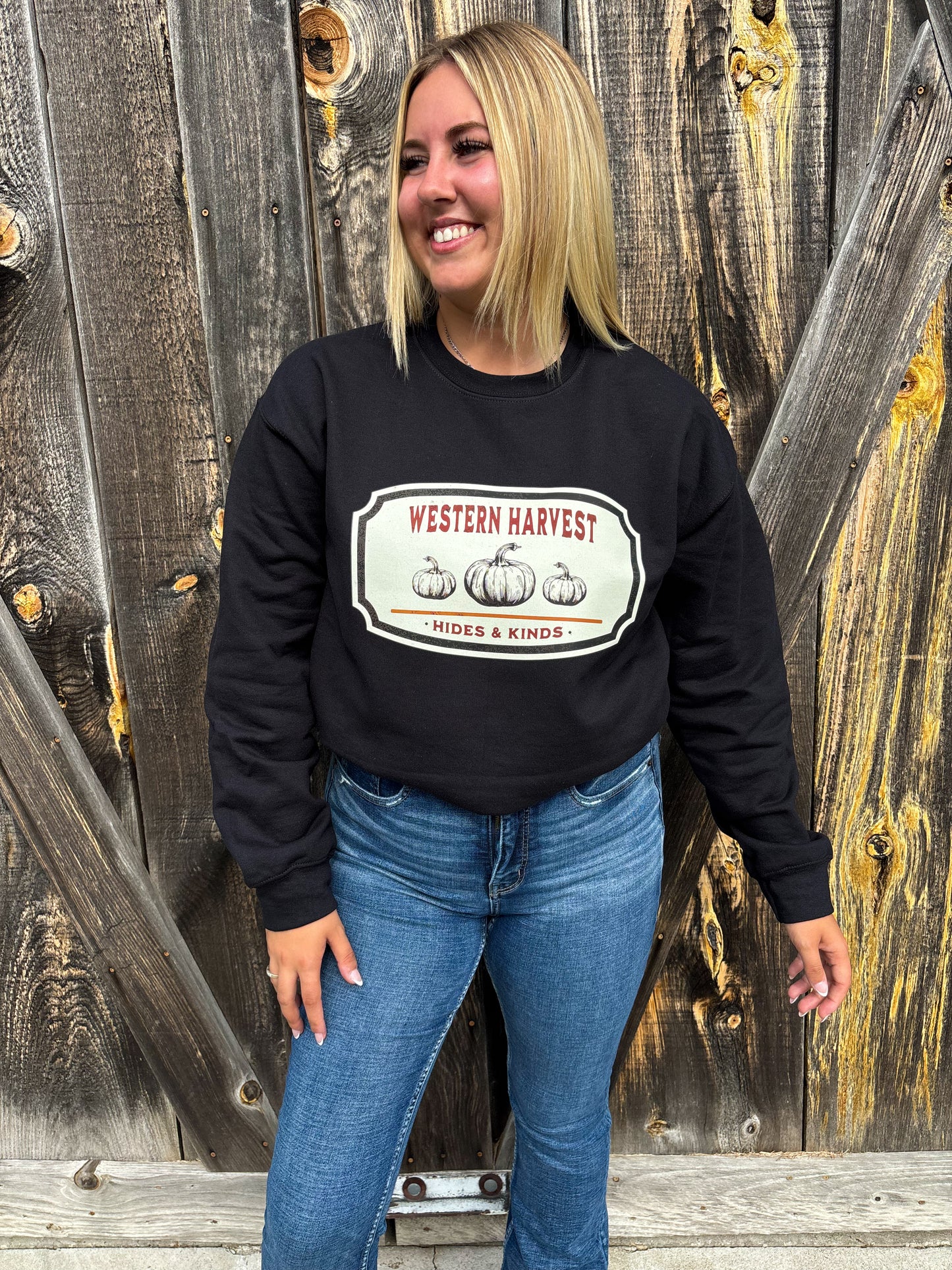 Full body shot of smiling model wearing Western Harvest crewneck by Hides & Kinds, standing in front of wood wall.
