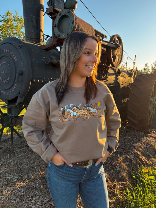 Model wearing the brown Wild Run Crewneck by Hides & Kinds with blue jeans, photographed outdoors beside vintage farm machinery.