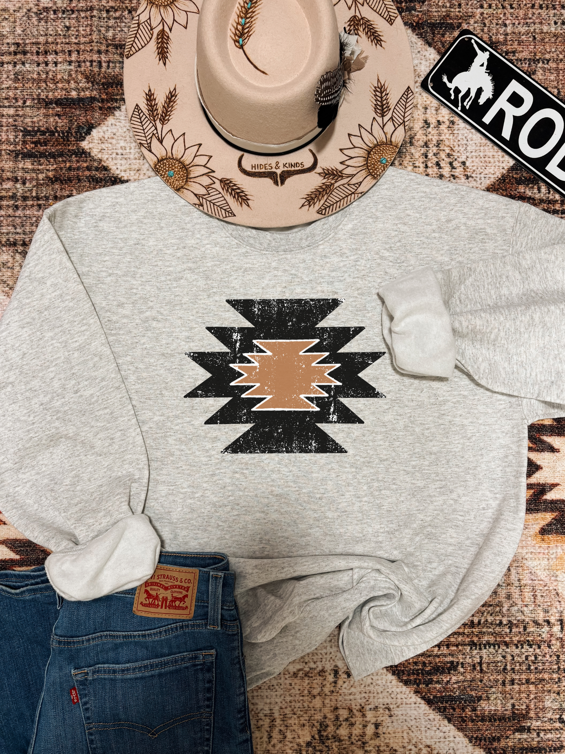 Flat lay of Aztec print crewneck in ash grey, styled with jeans, tan western hat, and a “Rodeo” sign on rustic background.
