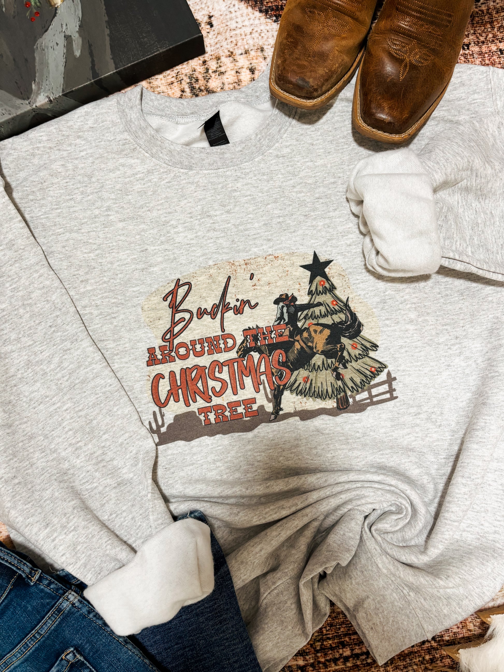 Ash grey crewneck featuring a bold western Christmas graphic of a cowboy riding a bull beside a decorated tree with a star on top. “Buckin’ Around the Christmas Tree” text in red. Styled flat lay with denim jeans and a rustic winter inspired backdrop.
