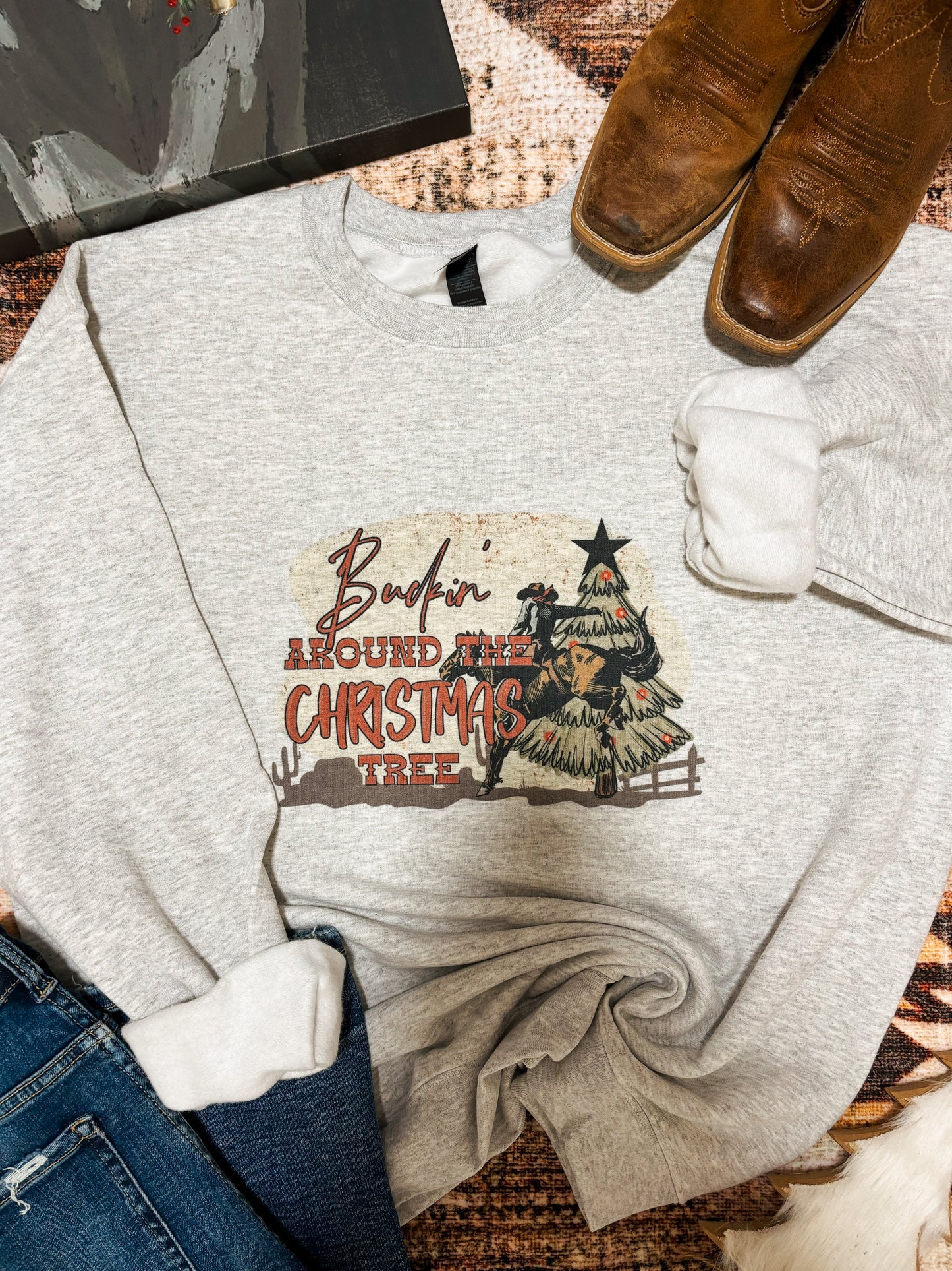 Flat lay of a western Christmas crewneck with “Buckin’ Around the Christmas Tree” graphic. Ash grey sweatshirt with cowboy riding a bull beside a decorated cactus-style tree. Styled with brown square-toe boots, denim jeans, and a cozy neutral background.