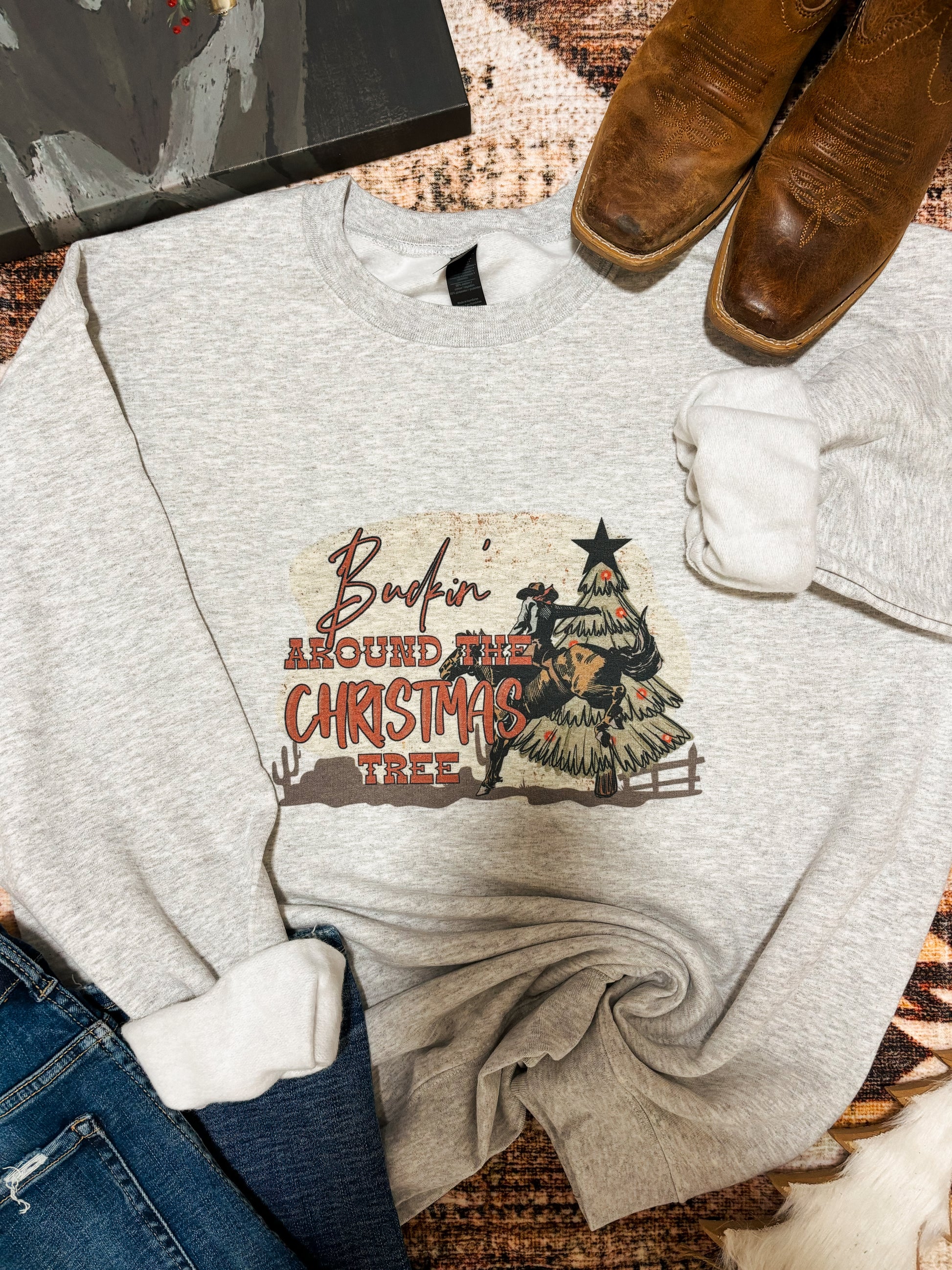 Flat lay of a western Christmas crewneck with “Buckin’ Around the Christmas Tree” graphic. Ash grey sweatshirt with cowboy riding a bull beside a decorated cactus-style tree. Styled with brown square-toe boots, denim jeans, and a cozy neutral background.