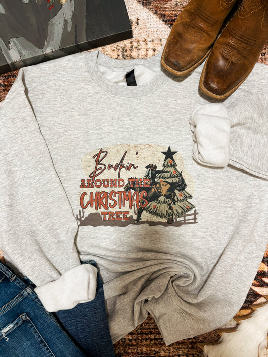 Flat lay of a western Christmas crewneck with “Buckin’ Around the Christmas Tree” graphic. Ash grey sweatshirt with cowboy riding a bull beside a decorated cactus-style tree. Styled with brown square-toe boots, denim jeans, and a cozy neutral background.