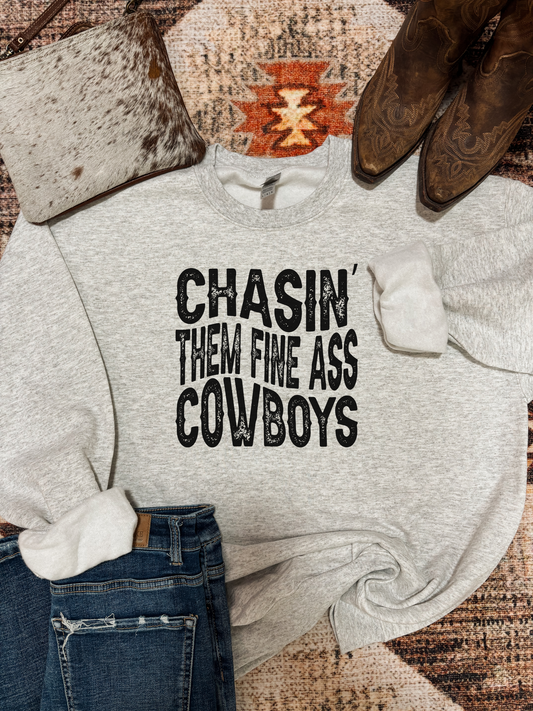 Heather grey crewneck with bold black western text that reads "Chasin' Them Fine Ass Cowboys," styled with jeans, cowboy boots, and a cowhide bag on a southwestern rug.