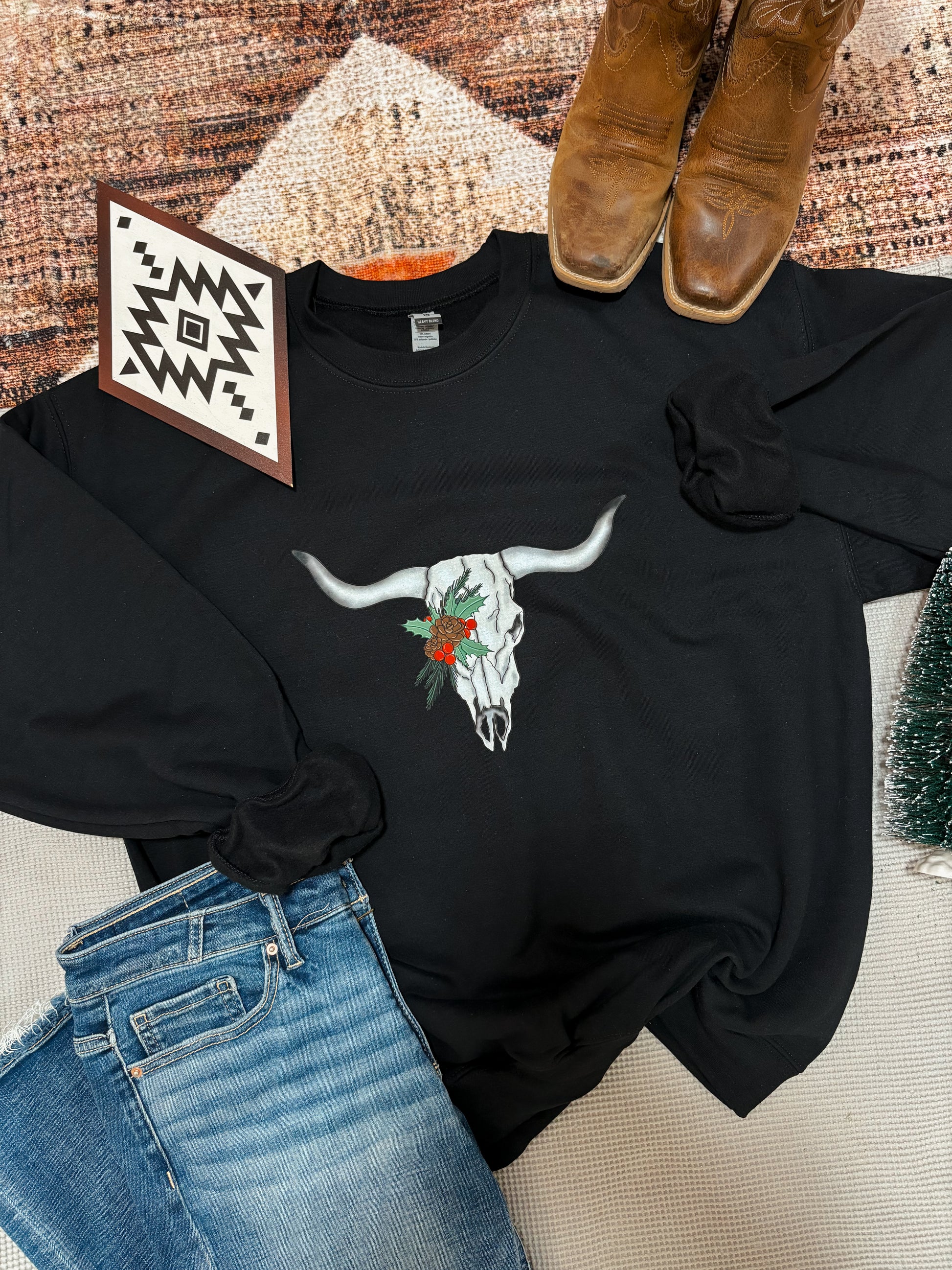 Western Christmas sweatshirt with a festive skull design, laid out beside cowboy boots, jeans, and a Southwestern boho print card.