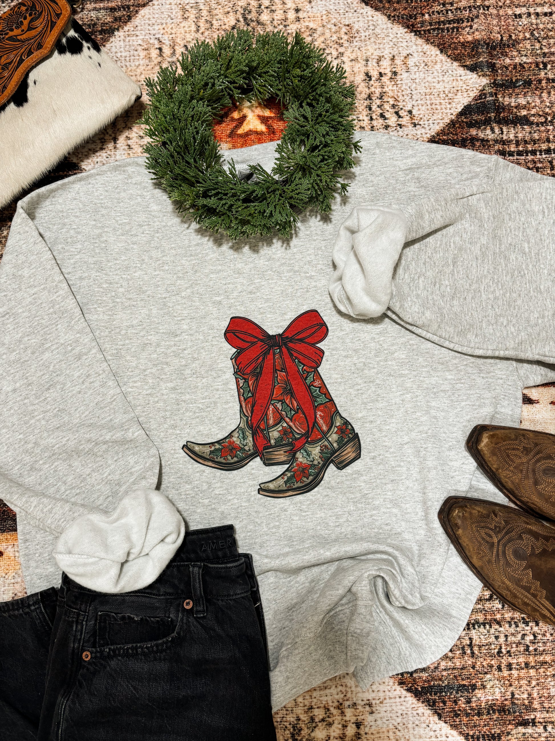 Flat lay of grey holiday crewneck featuring red western boots wrapped in a bow, styled with wreath and western accessories