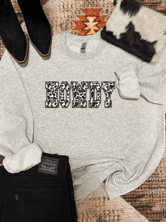Flat lay of the Cowprint Howdy Crewneck in athletic grey with bold cow print lettering, styled with black jeans, cowhide clutch, and suede boots.