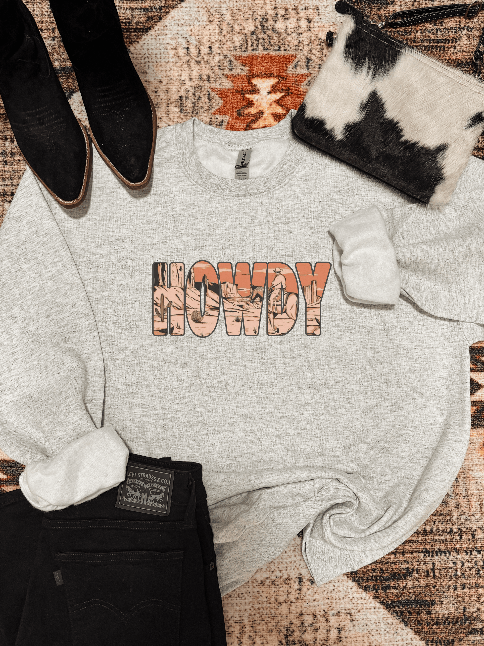Heather grey crewneck with desert themed "Howdy" graphic, styled with black jeans, boots, and cowhide bag