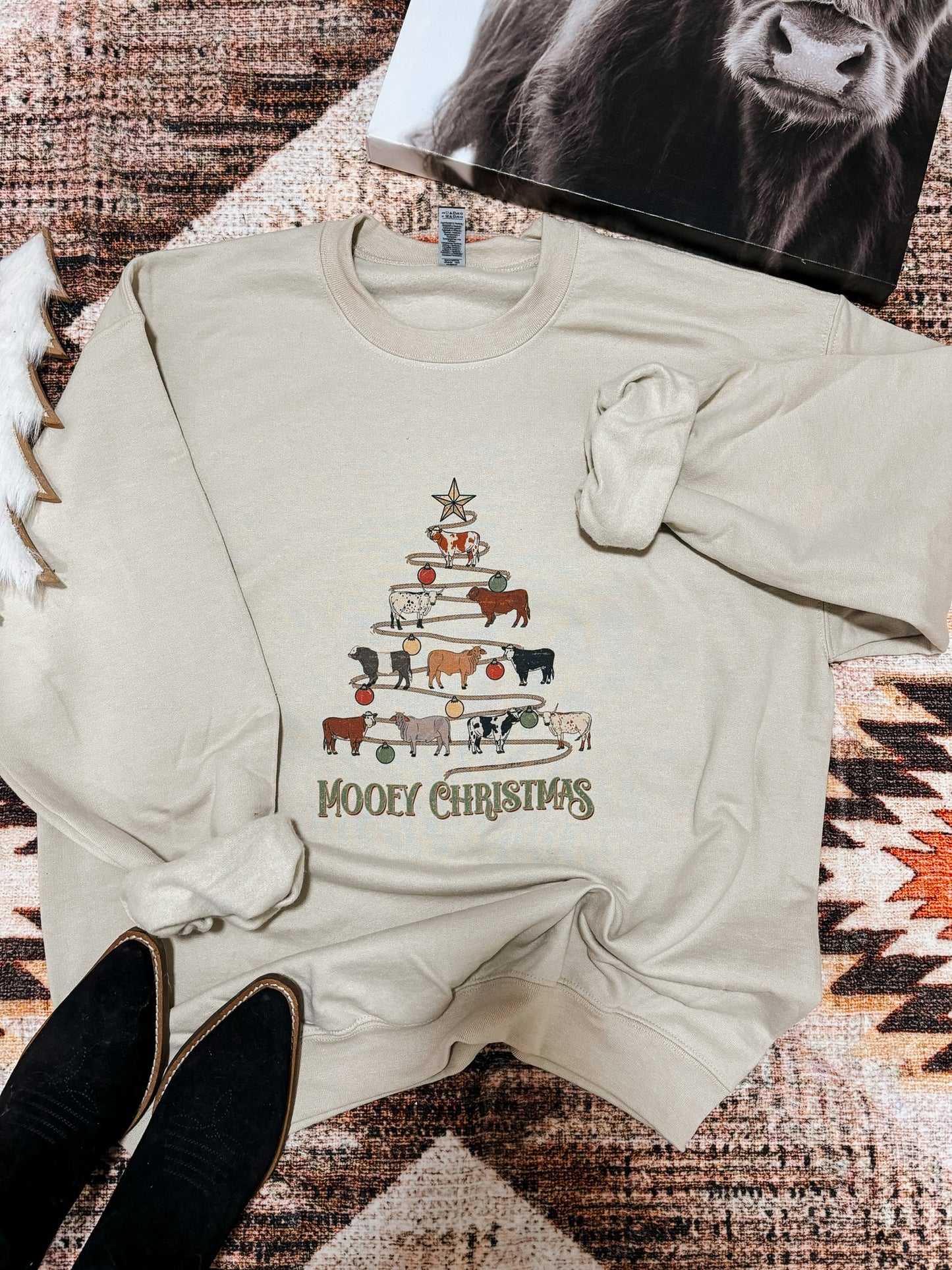Western holiday sweatshirt with a playful cow tree graphic and "Mooey Christmas" wording, photographed flatlay with boots and a western art print.