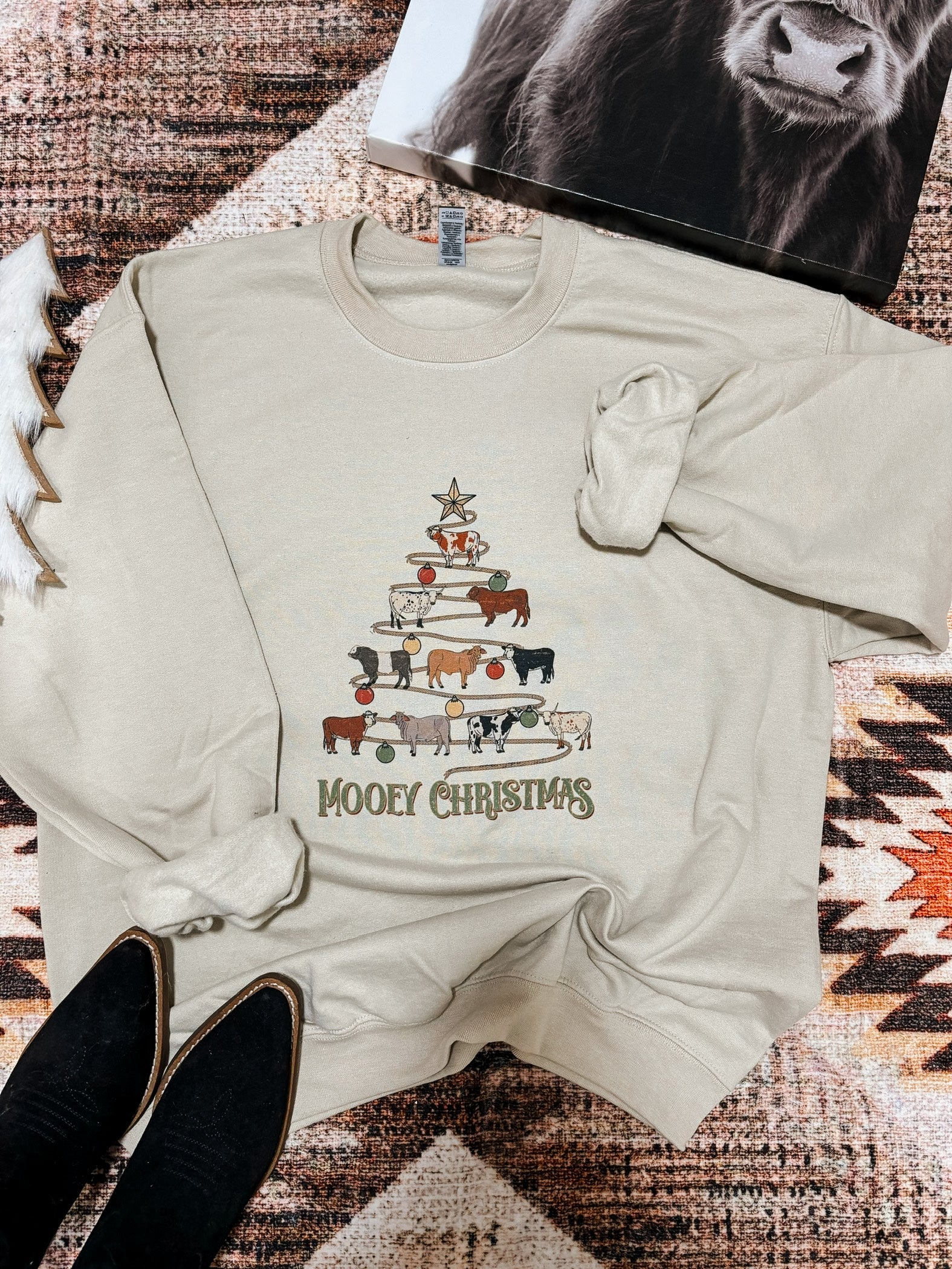 Western holiday sweatshirt with a playful cow tree graphic and "Mooey Christmas" wording, photographed flatlay with boots and a western art print.