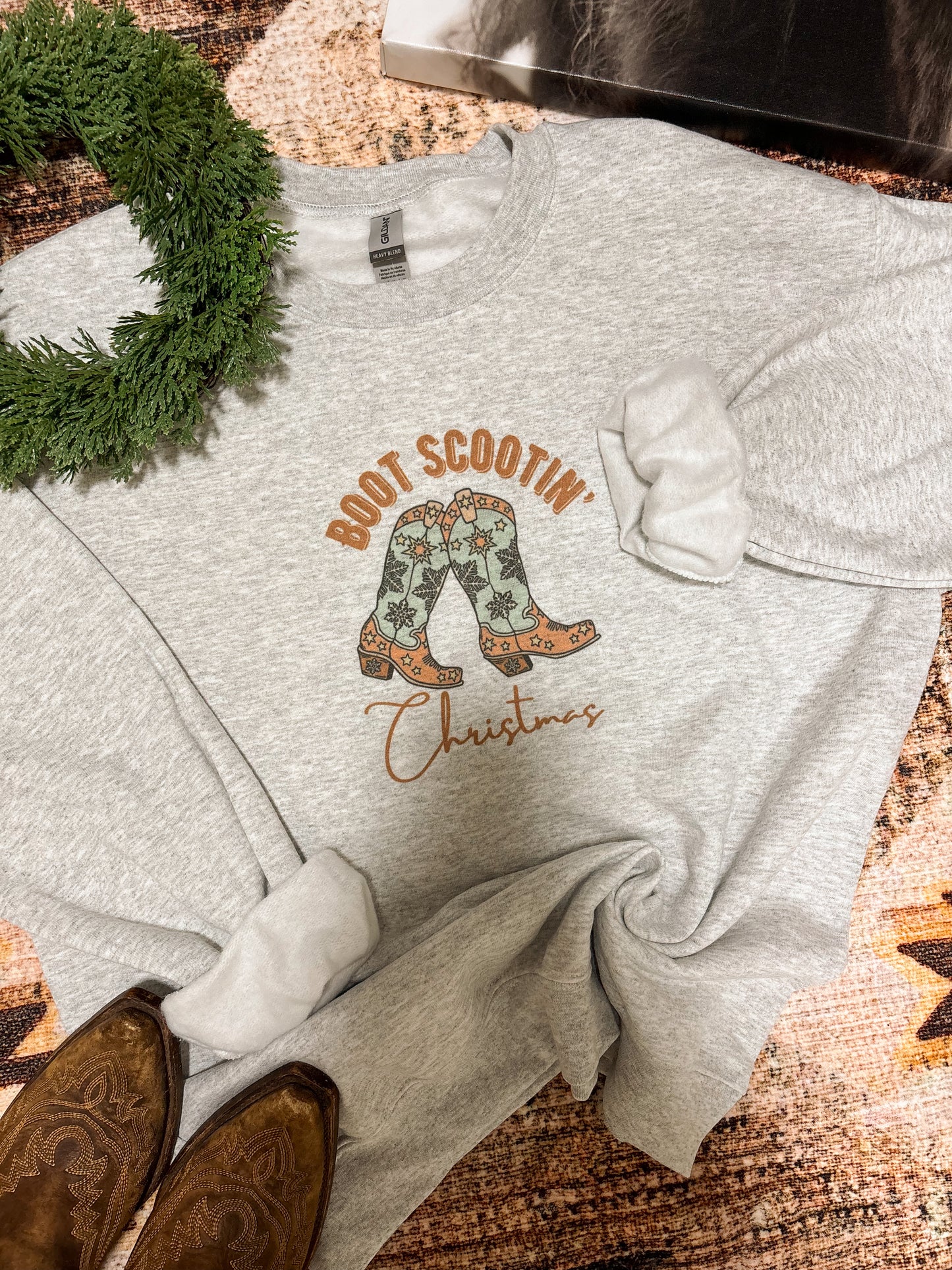 Christmas cowgirl sweatshirt with boot graphic and wreath accent, laid flat on western rug