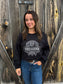 Smiling woman in the black Hides & Kinds Outback Crewneck against barnwood backdrop