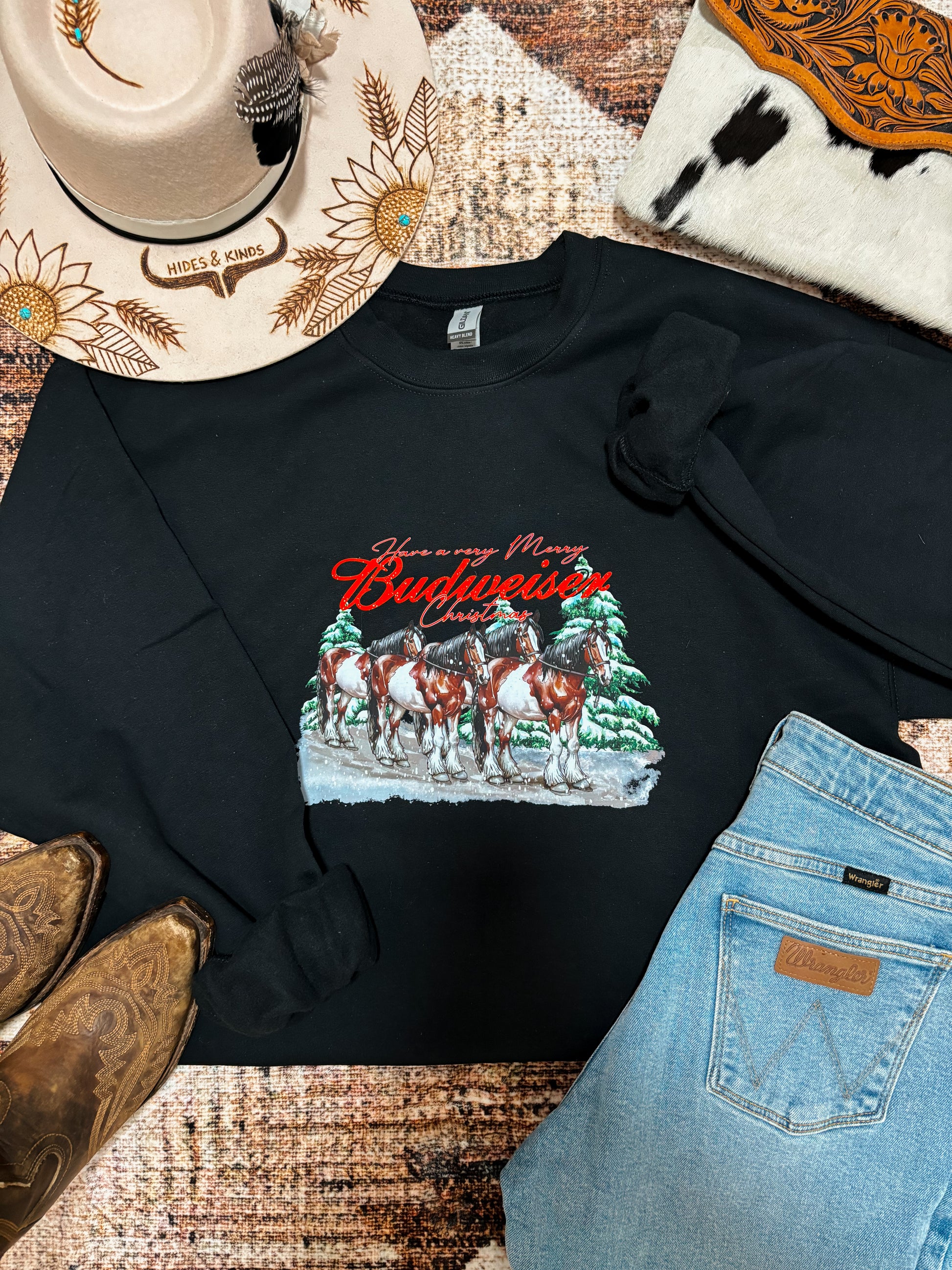 Western flat lay of black Budweiser Christmas crewneck styled with cowboy boots, denim, and Hides & Kinds sunflower hat.