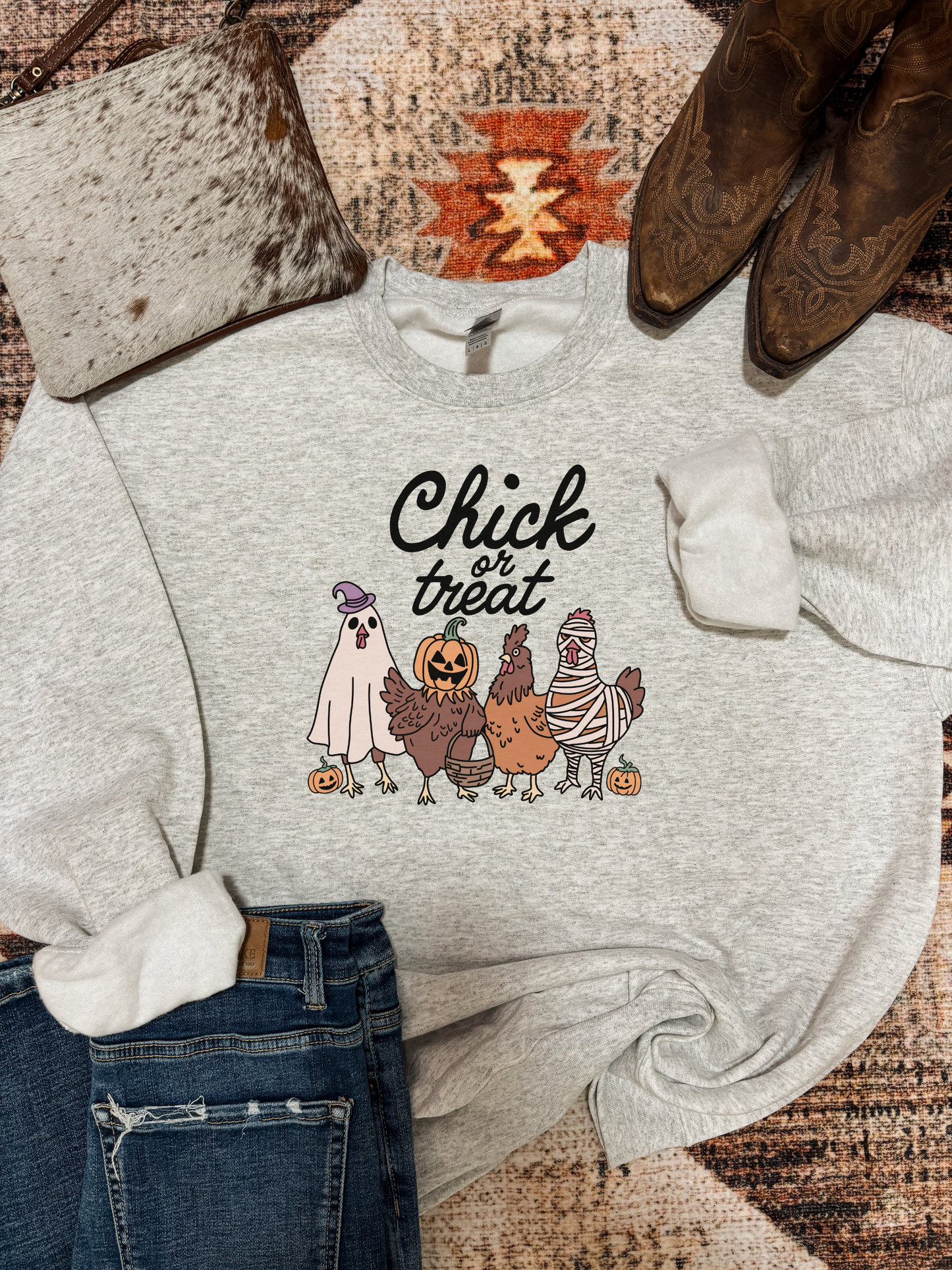 Grey crewneck featuring four cartoon chickens dressed in Halloween costumes with pumpkins and “Chick or Treat” text, styled with jeans, cowhide purse, and western boots